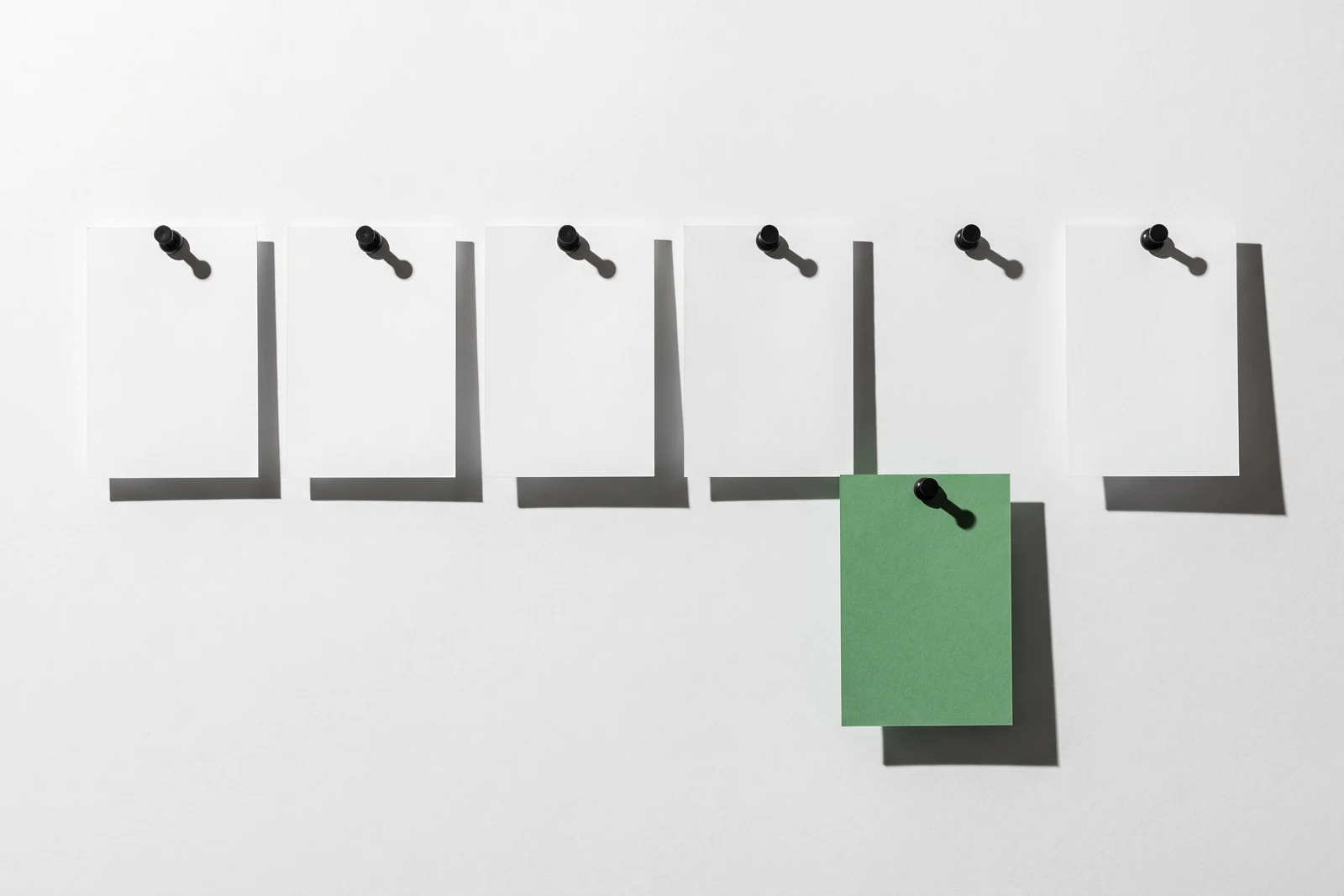 Six cartes de papier blanc punaisées en ligne sur une surface blanche, la cinquième, en papier vert, glissée d’un cran vers le bas.