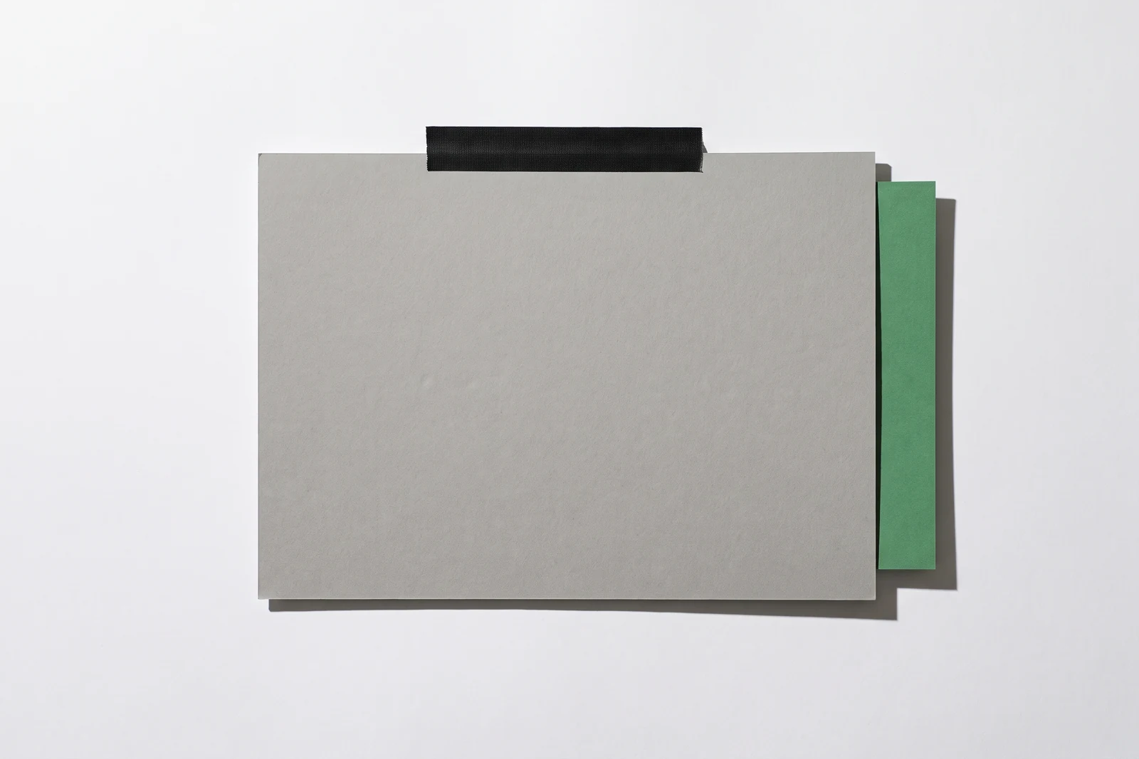 Une plaque de carton gris tenue par un ruban noir contre une surface blanche, une bande de papier vert dépassant de son bord droit.