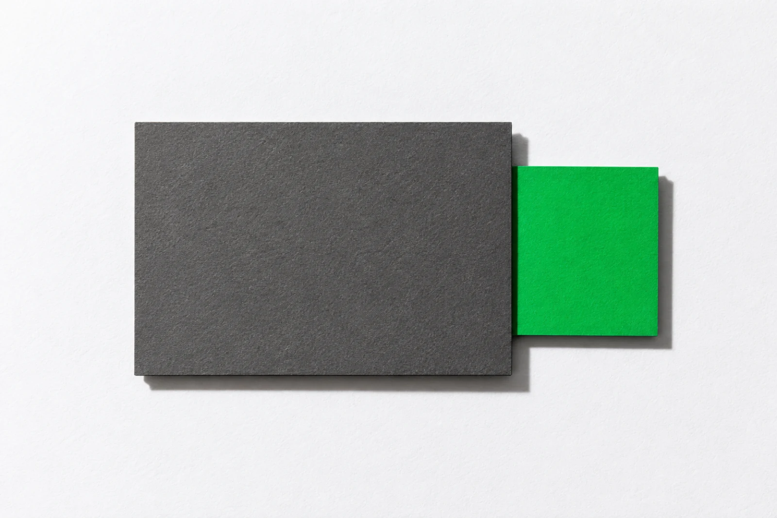Une petite carte de papier vert glissée à moitié sous une épaisse carte de carton gris, posées à plat sur un fond blanc, ombres dures projetées vers le bas à droite.