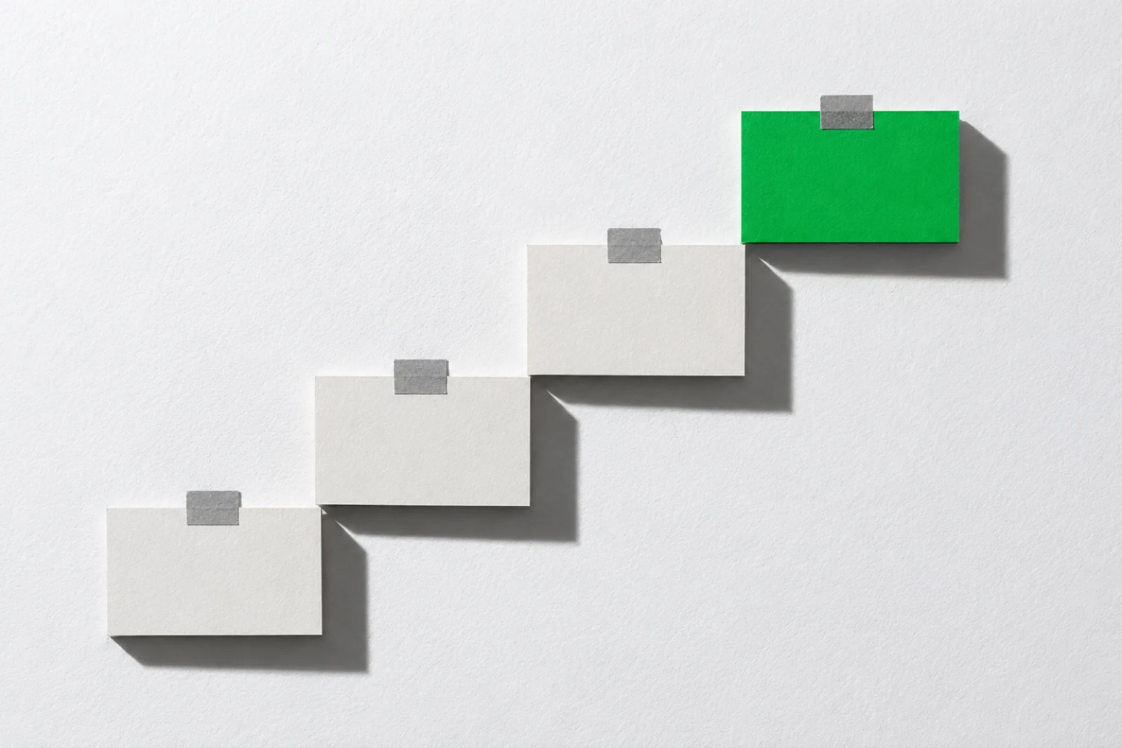 Quatre cartes de papier fixées au mur en escalier par de petites bandes de ruban gris, la marche du haut en vert, ombres dures projetées vers le bas à droite.
