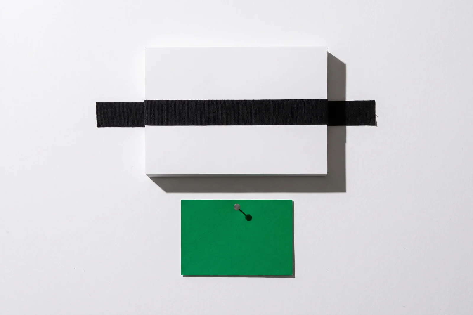 Un bloc épais de cartes de papier blanc maintenu contre une surface verticale par une bande de toile noire tendue en travers, et une carte de papier vert punaisée juste en dessous, ombres dures vers le bas à droite.