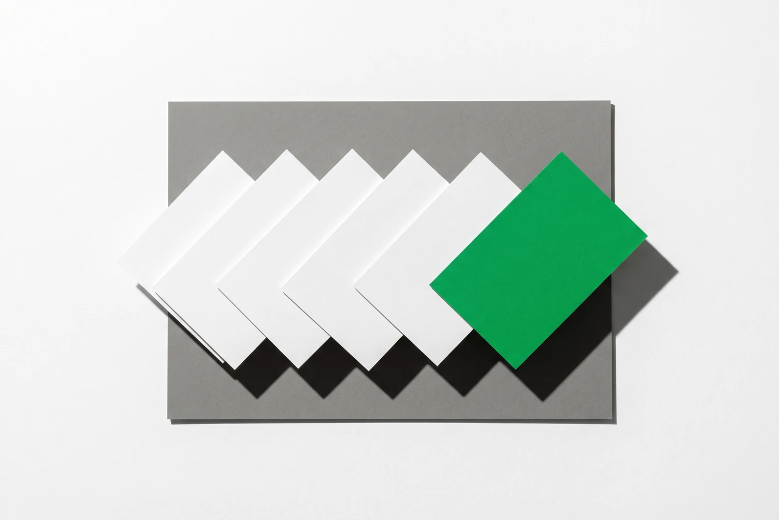 Six cartes de papier posées en ligne sur une plaque de carton gris, chacune décalée de la précédente du même écart, la dernière en papier vert posée par-dessus, ombres dures vers le bas à droite.