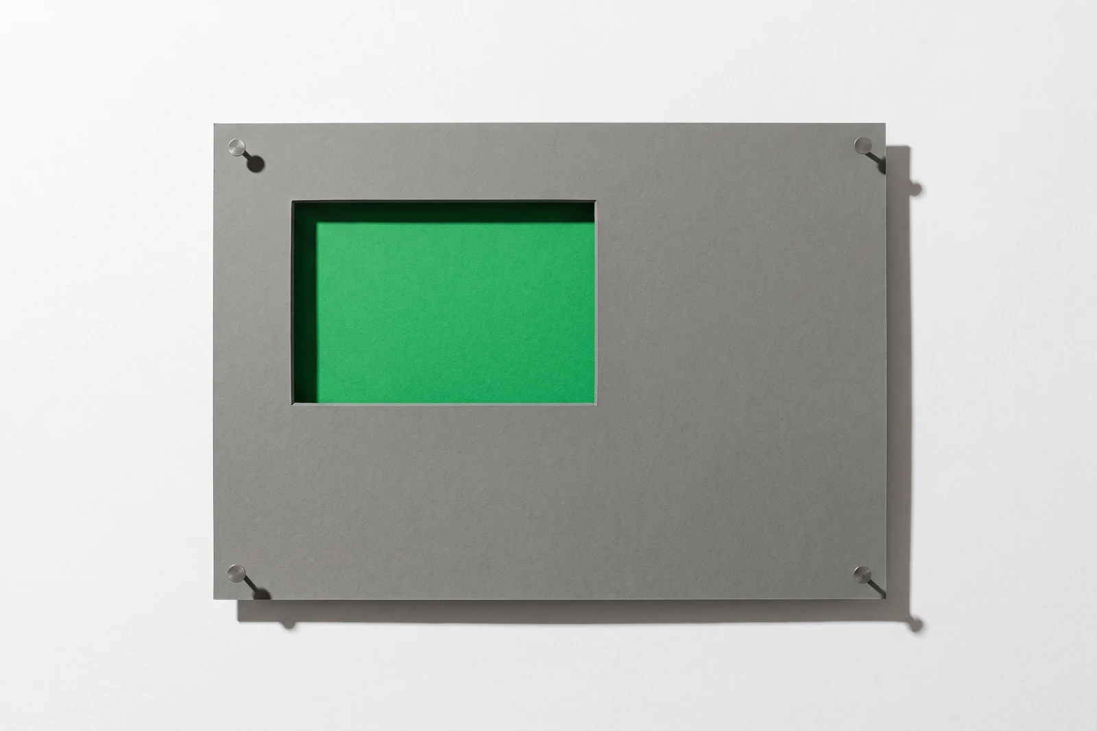 Une plaque de carton gris punaisée aux quatre coins sur une surface blanche, percée d’une fenêtre rectangulaire qui laisse voir une carte de papier vert posée derrière, ombre fine dans la fenêtre vers le bas à droite.