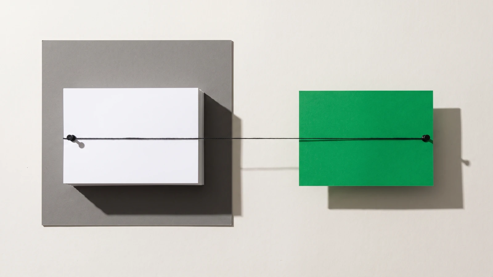 Une pile épaisse de cartes de papier blanc posée sur une plaque de carton gris et une carte de papier vert posée à distance, reliées par un seul fil de coton noir tendu entre deux punaises, ombres dures projetées vers le bas à droite.