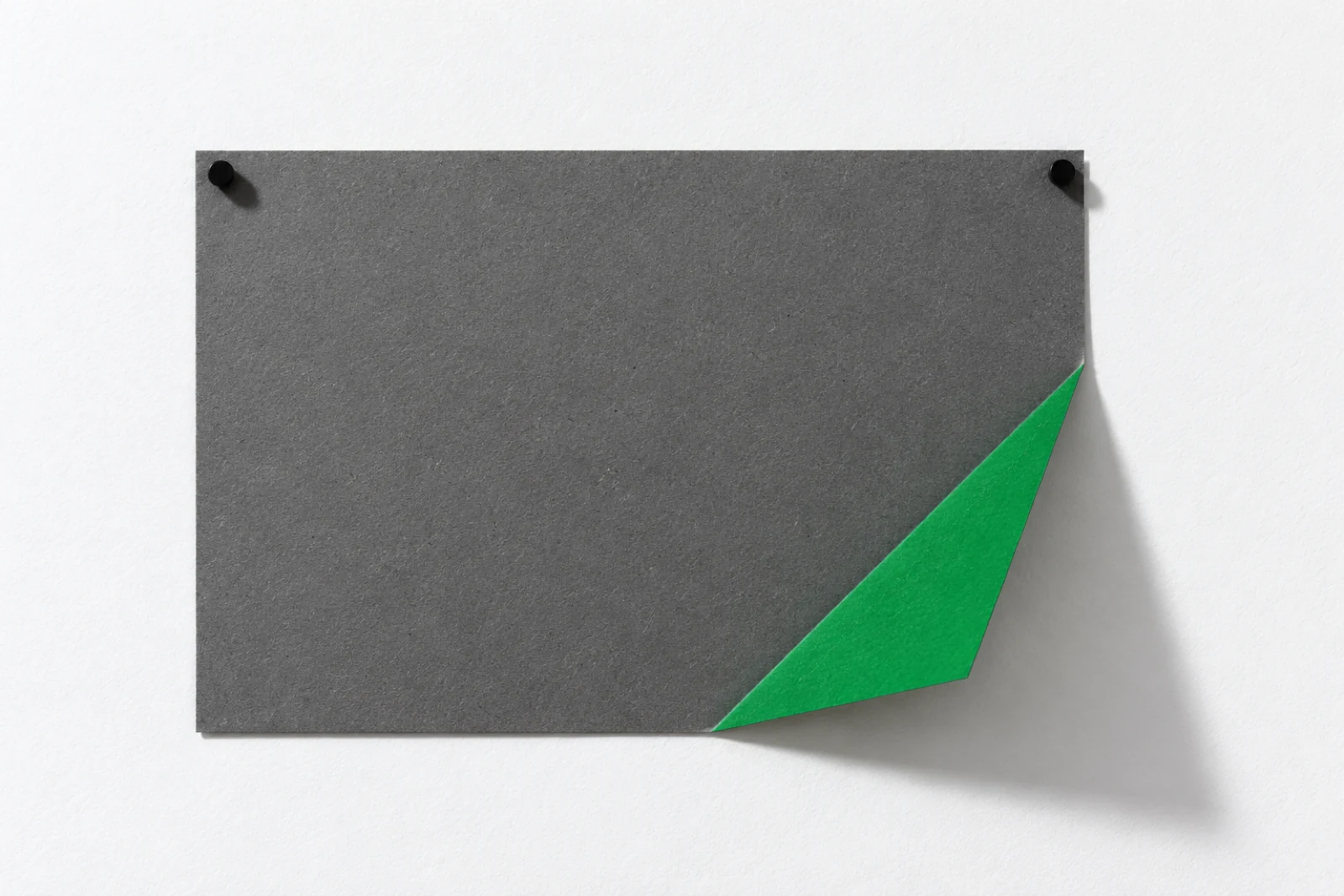 Une feuille de carton gris punaisée sur un mur blanc, son coin inférieur droit replié en triangle qui montre un revers vert, ombre nette portée vers le bas à droite.