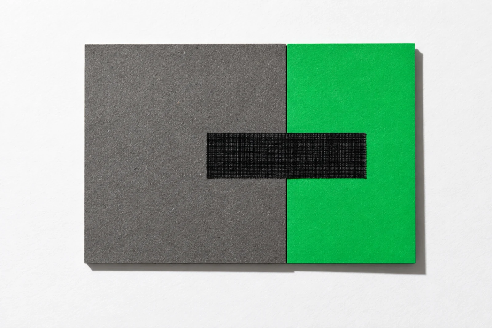Une plaque de carton gris et une carte verte posées bord à bord sur un fond blanc, réunies par une seule bande de toile noire collée en travers de la jointure.