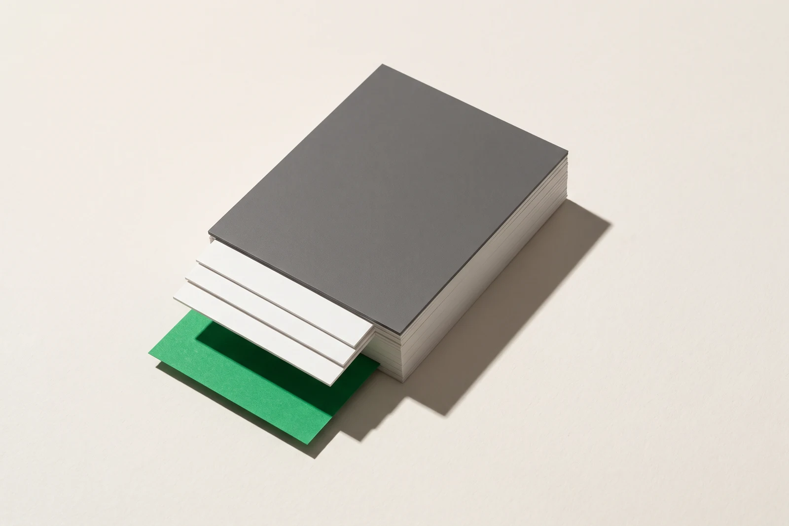 Une pile épaisse de cartes de papier vierge, coiffée d'un carton gris mat ; les quatre cartes du haut dépassent franchement du carton, et une feuille verte glisse dessous.