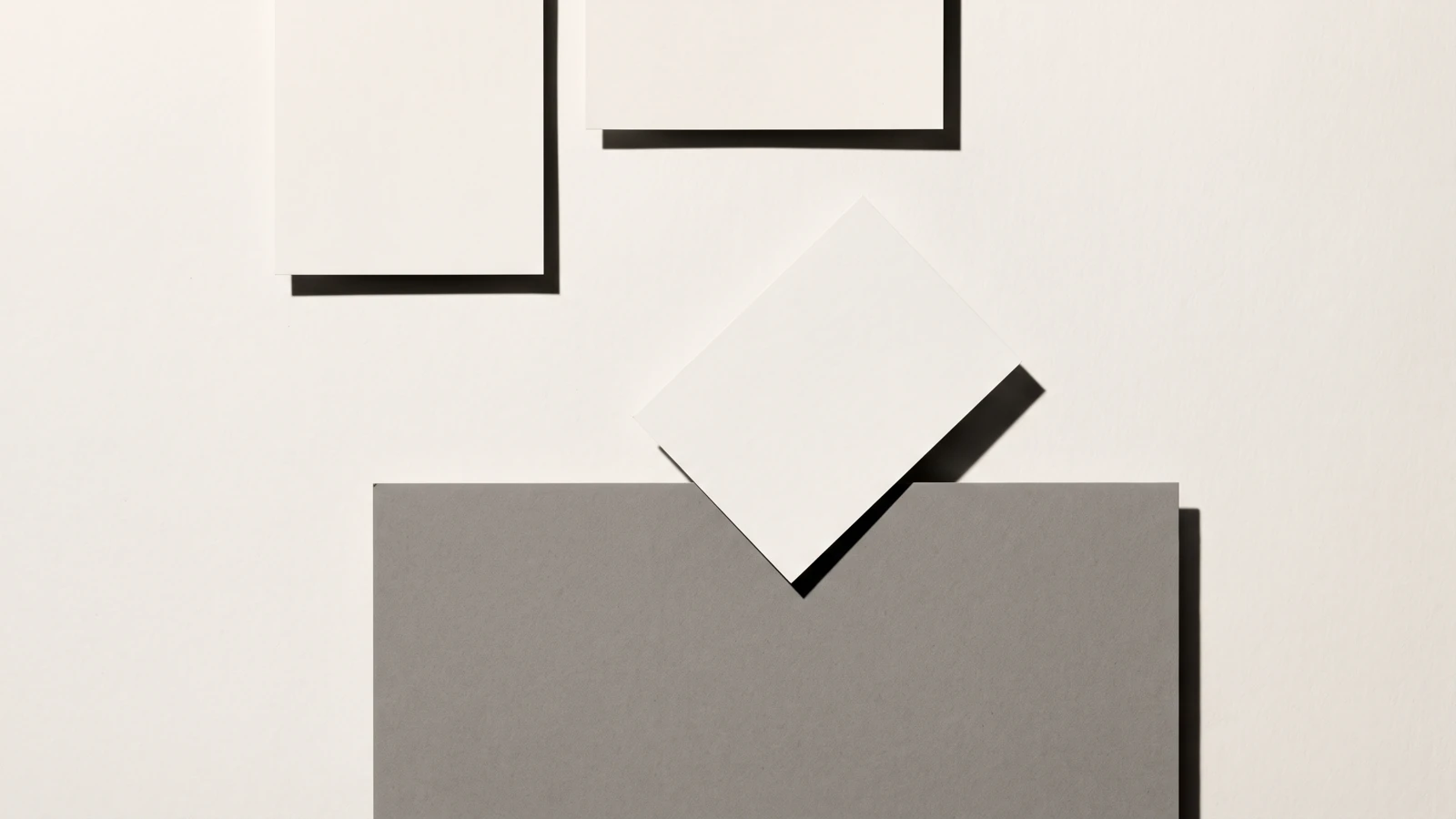Une carte de papier en équilibre sur le bord d’une plaque de carton gris, ombres dures projetées vers le bas à droite.