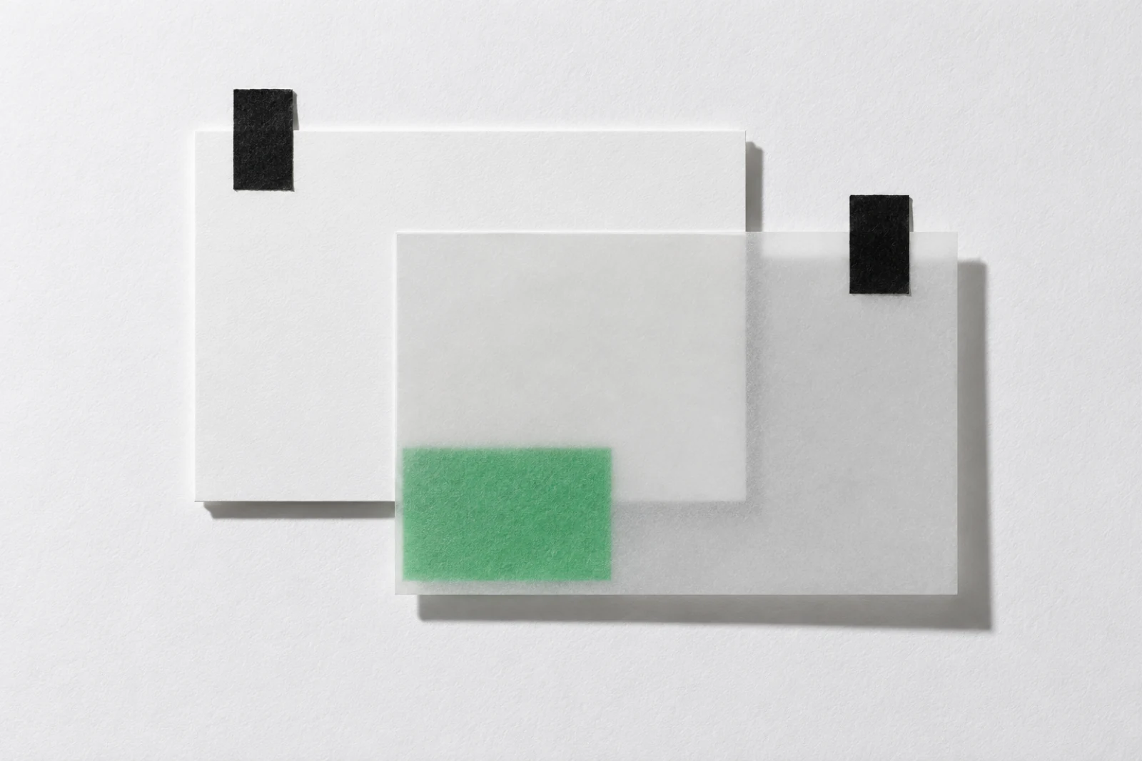 Une carte blanche scotchée par deux bandes noires, recouverte en décalage d'une feuille de papier calque qui laisse voir un petit rectangle vert glissé dessous.
