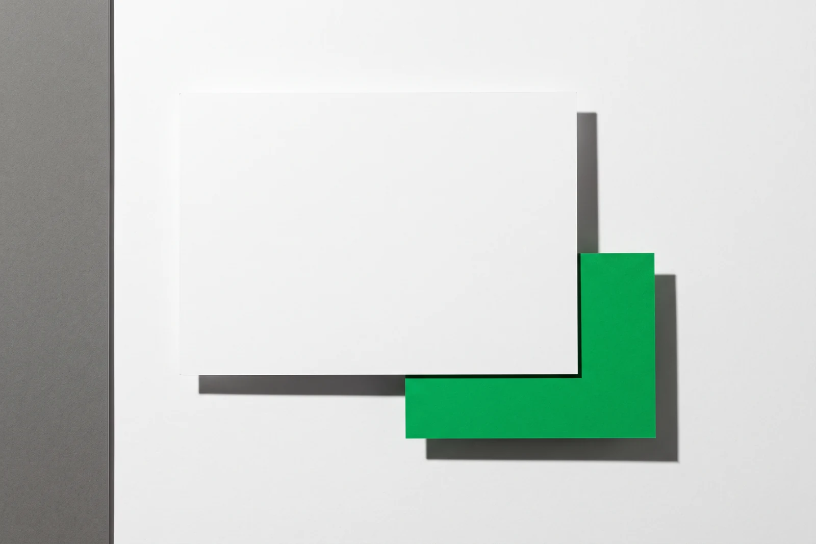 Une carte de papier vierge glissée sous une plus grande, dont un angle teinté de vert dépasse en bas à droite, ombre nette portée.