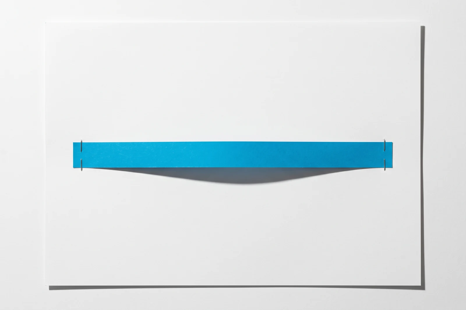 Une bande de papier cyan tendue entre deux agrafes plates, soulevée en son milieu, avec son ombre nette en dessous.