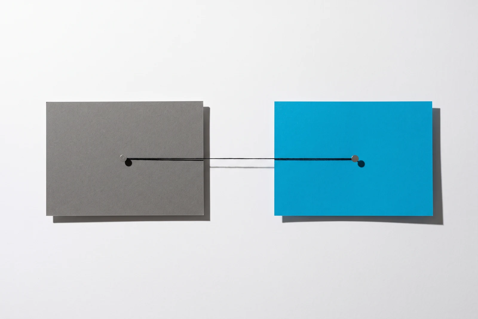 Deux rectangles épinglés côte à côte, l'un en carton gris, l'autre en papier cyan, reliés par un fil de coton noir tendu à l'horizontale.