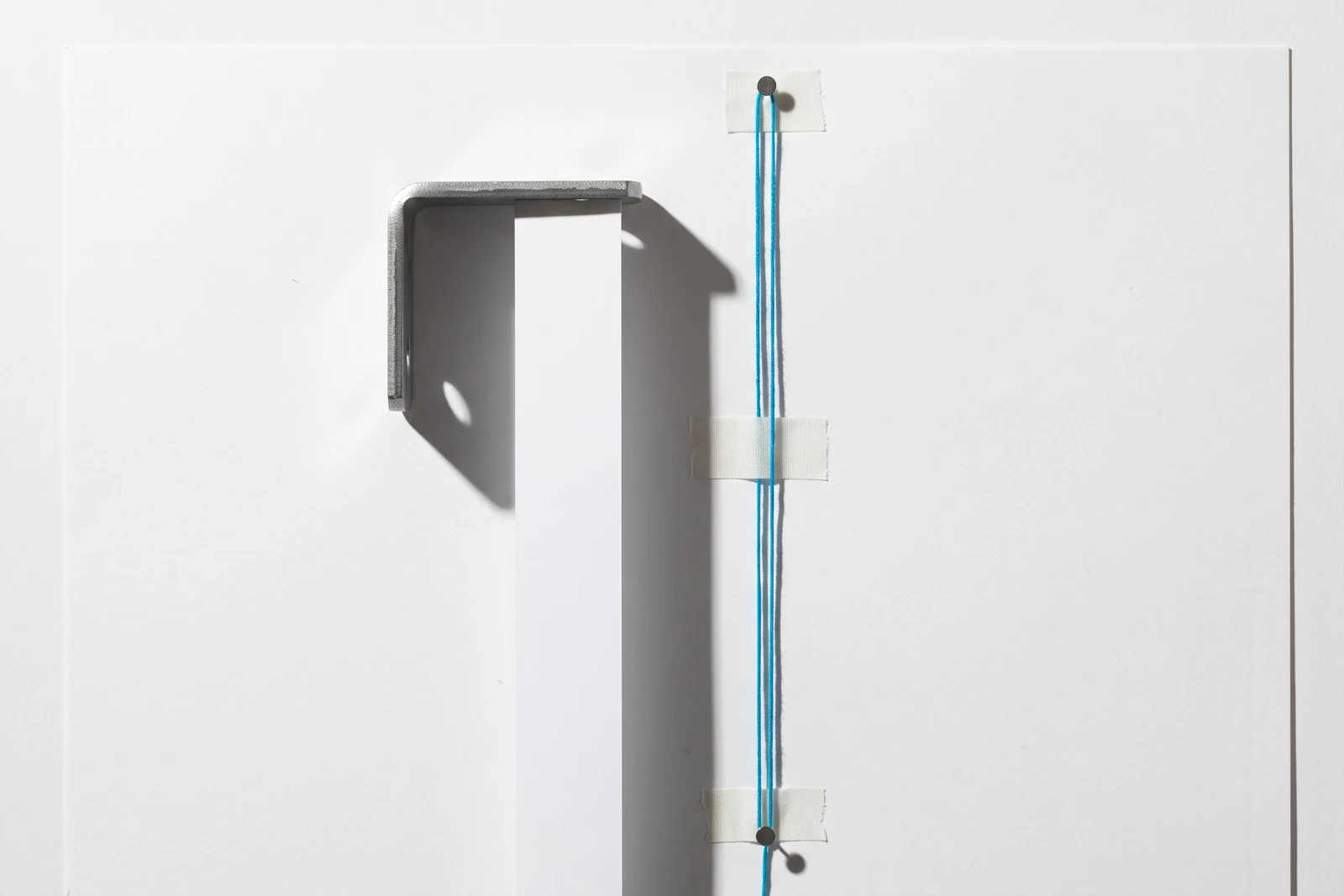Une bande de papier venant buter contre le bras d’une équerre d’acier mat, ombres dures projetées vers le bas à droite.