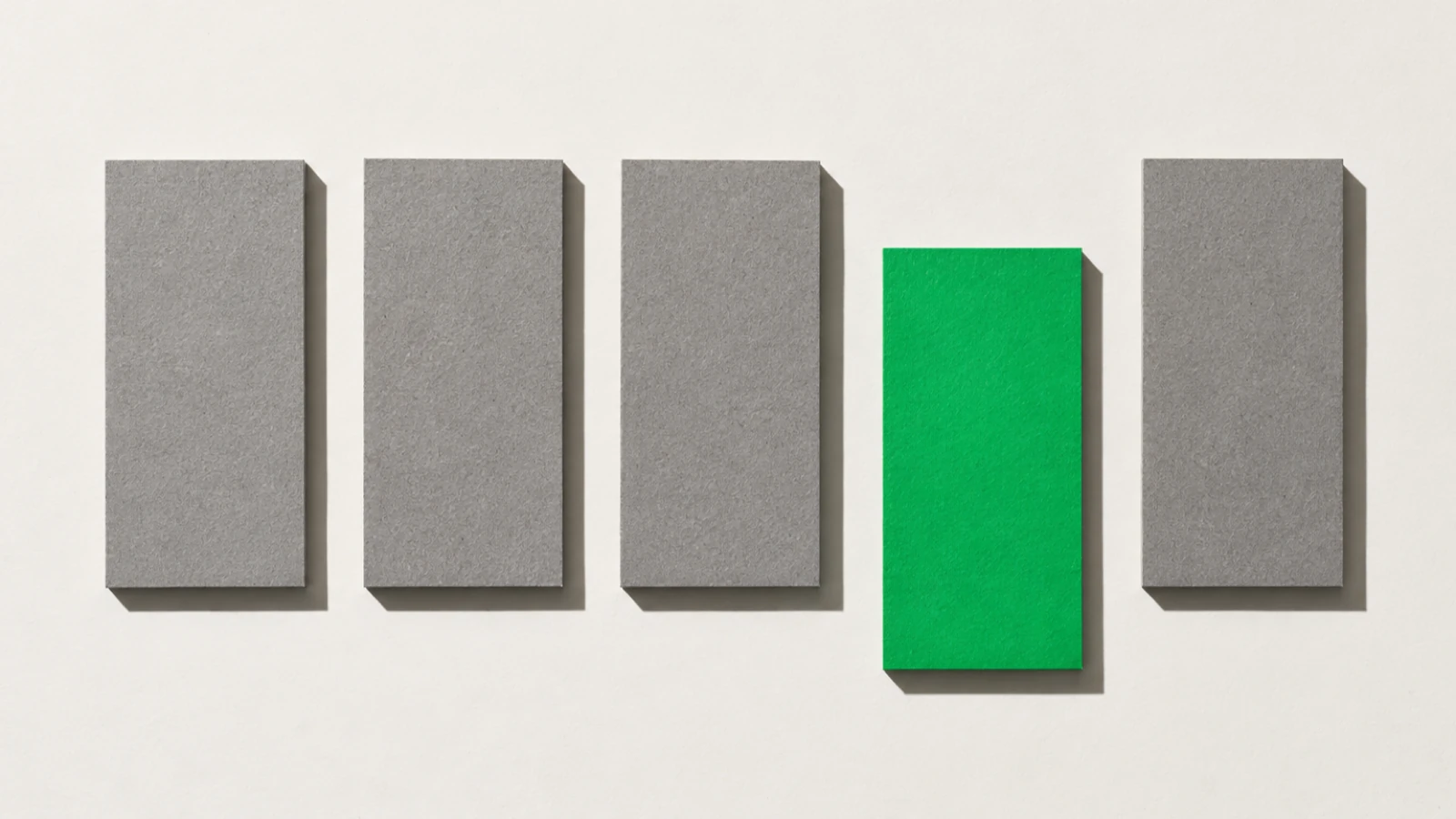 Vue de dessus, sur un fond de papier blanc cassé, cinq rectangles alignés à intervalles réguliers : quatre en carton gris et un en papier vert, décalé plus bas que les autres ; ombres dures vers le bas à droite.