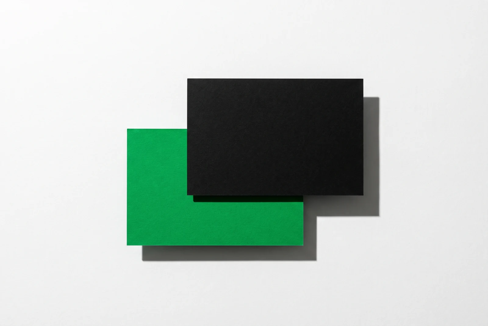 Deux cartes de papier épais sur fond blanc, une carte noire posée à plat et une carte verte glissée à moitié dessous par la gauche, ombre nette vers le bas à droite.