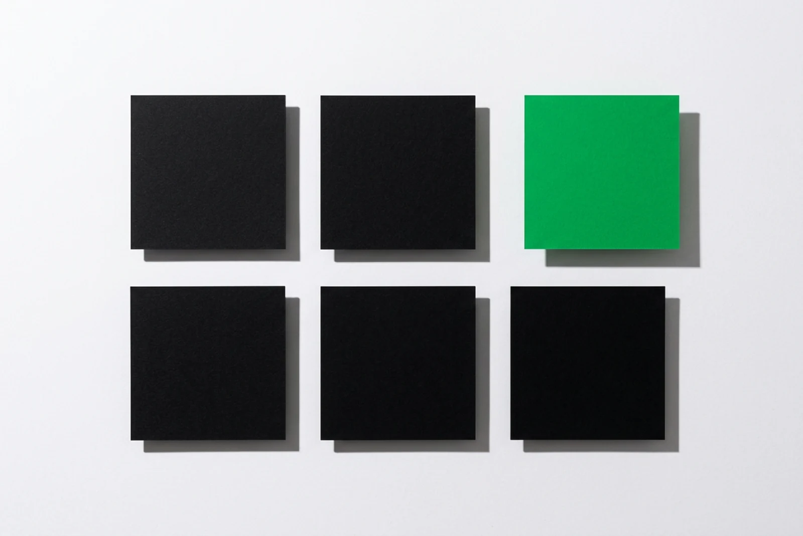 Six cartes carrées de papier posées en grille de deux rangées sur fond blanc, cinq noires et une verte décalée vers la droite hors de l’alignement, ombres dures vers le bas à droite.