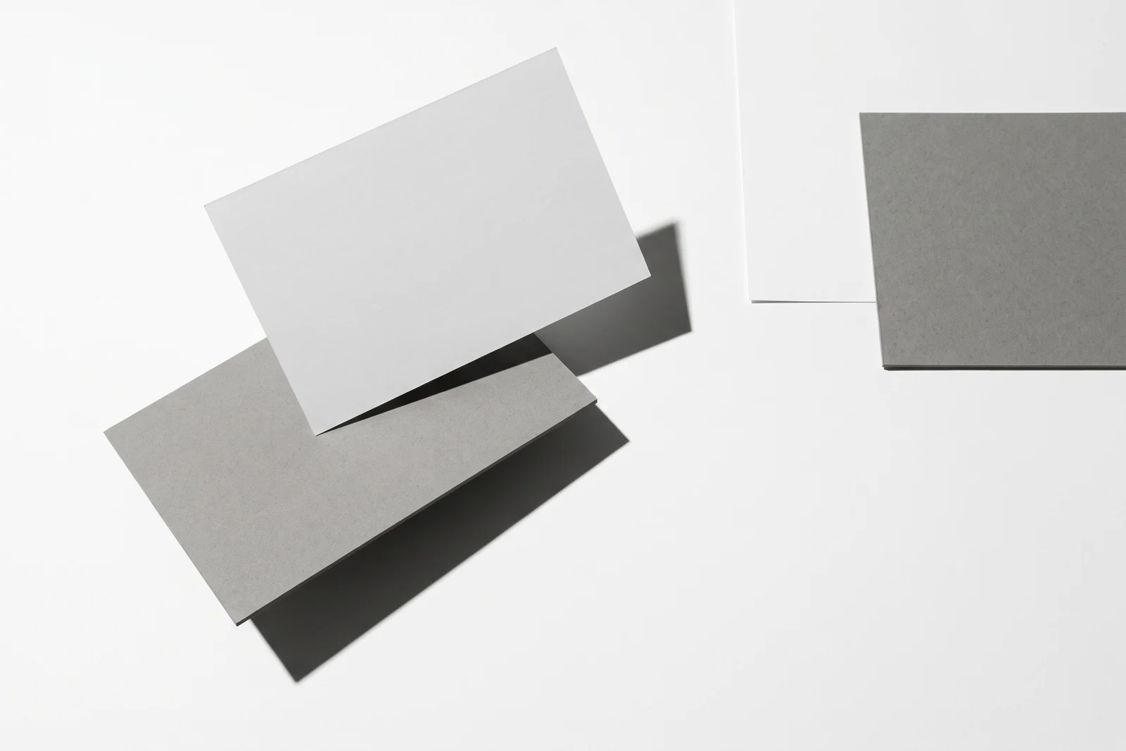 Une carte de papier en équilibre sur le bord d’une plaque de carton gris, ombres dures projetées vers le bas à droite.