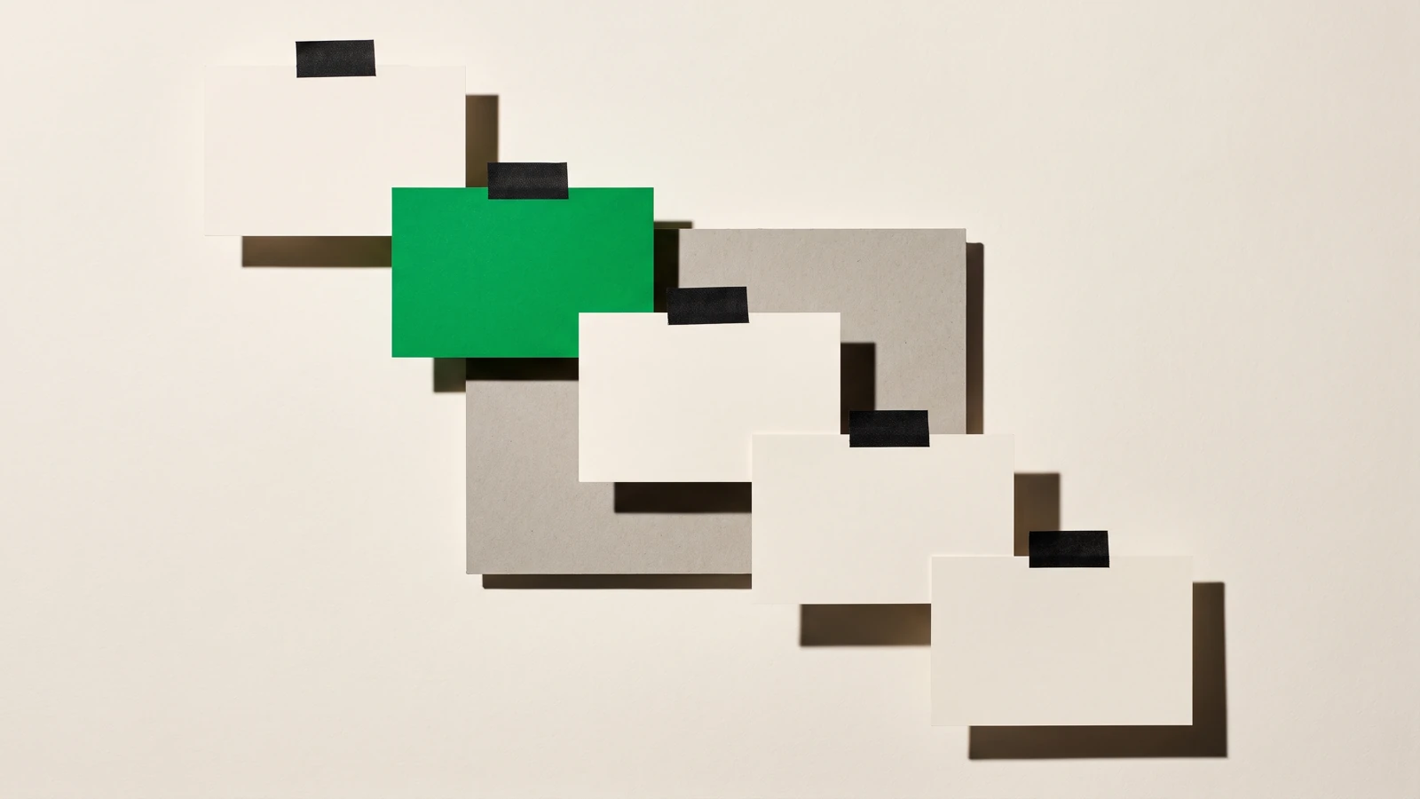 Cinq cartes de papier vierge scotchées en escalier descendant contre une surface verticale, la deuxième en papier vert, sur un fond de carton gris, ombres dures projetées vers le bas à droite.
