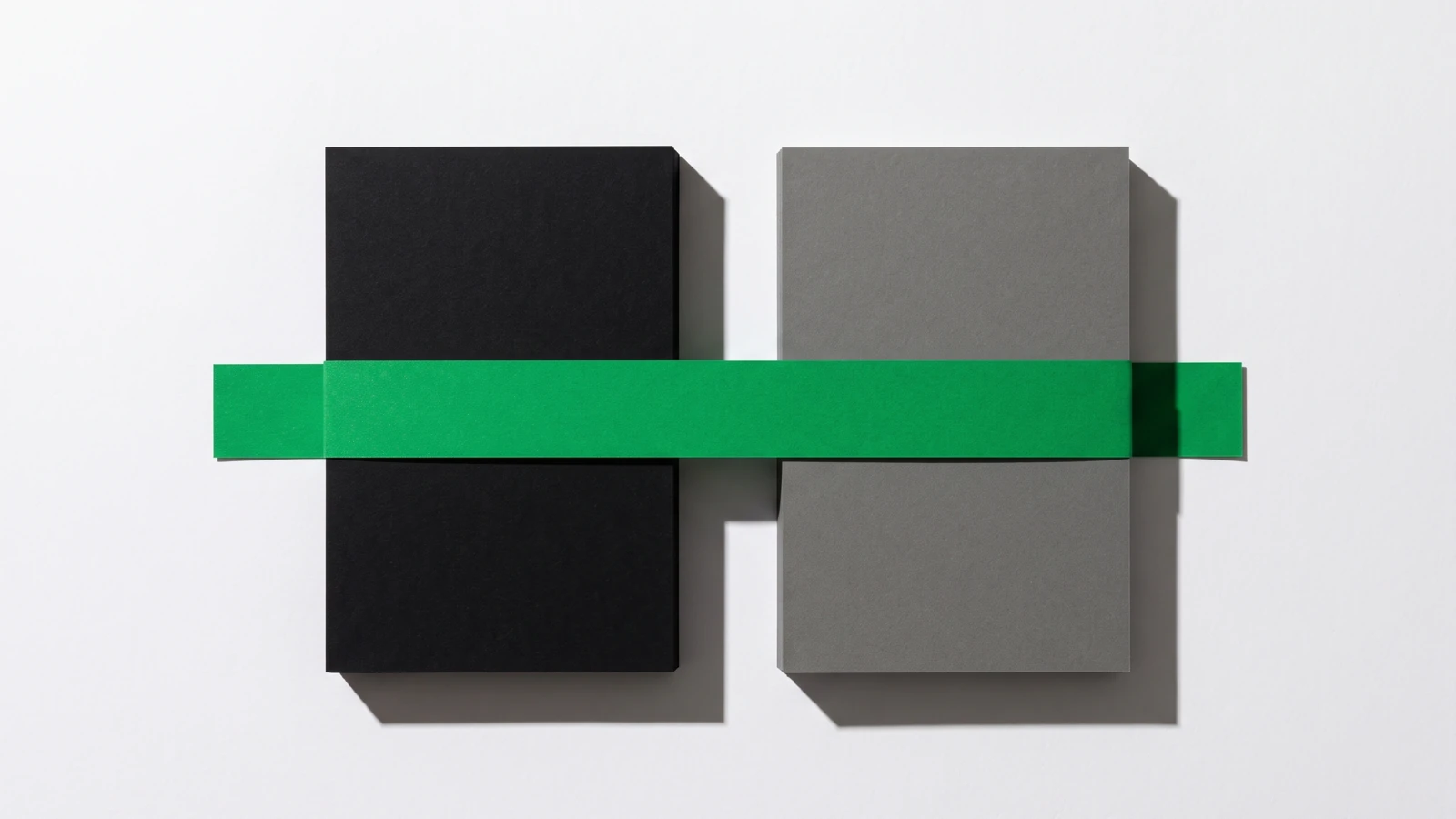 Deux piles de cartes de papier, une noire et une grise, posées côte à côte sur fond blanc et tenues ensemble par une seule bande de papier vert posée en travers, ombres nettes vers le bas à droite