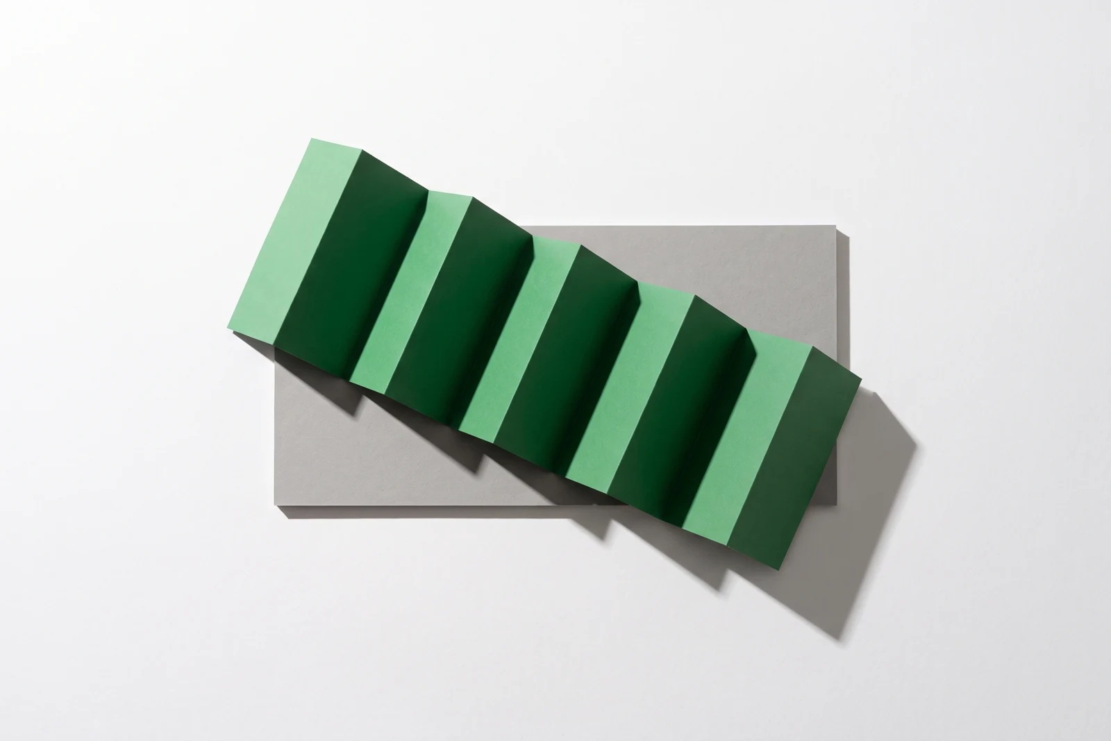 Une bande de papier vert pliée en accordéon de cinq panneaux, posée sur un rectangle de carton gris.