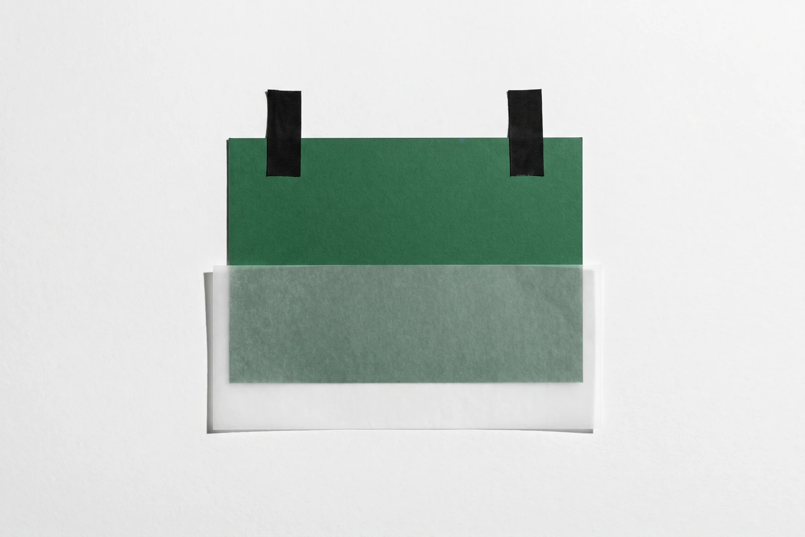 Un rectangle de papier vert scotché contre une surface blanche, sa moitié basse recouverte d’une feuille de calque cristal.