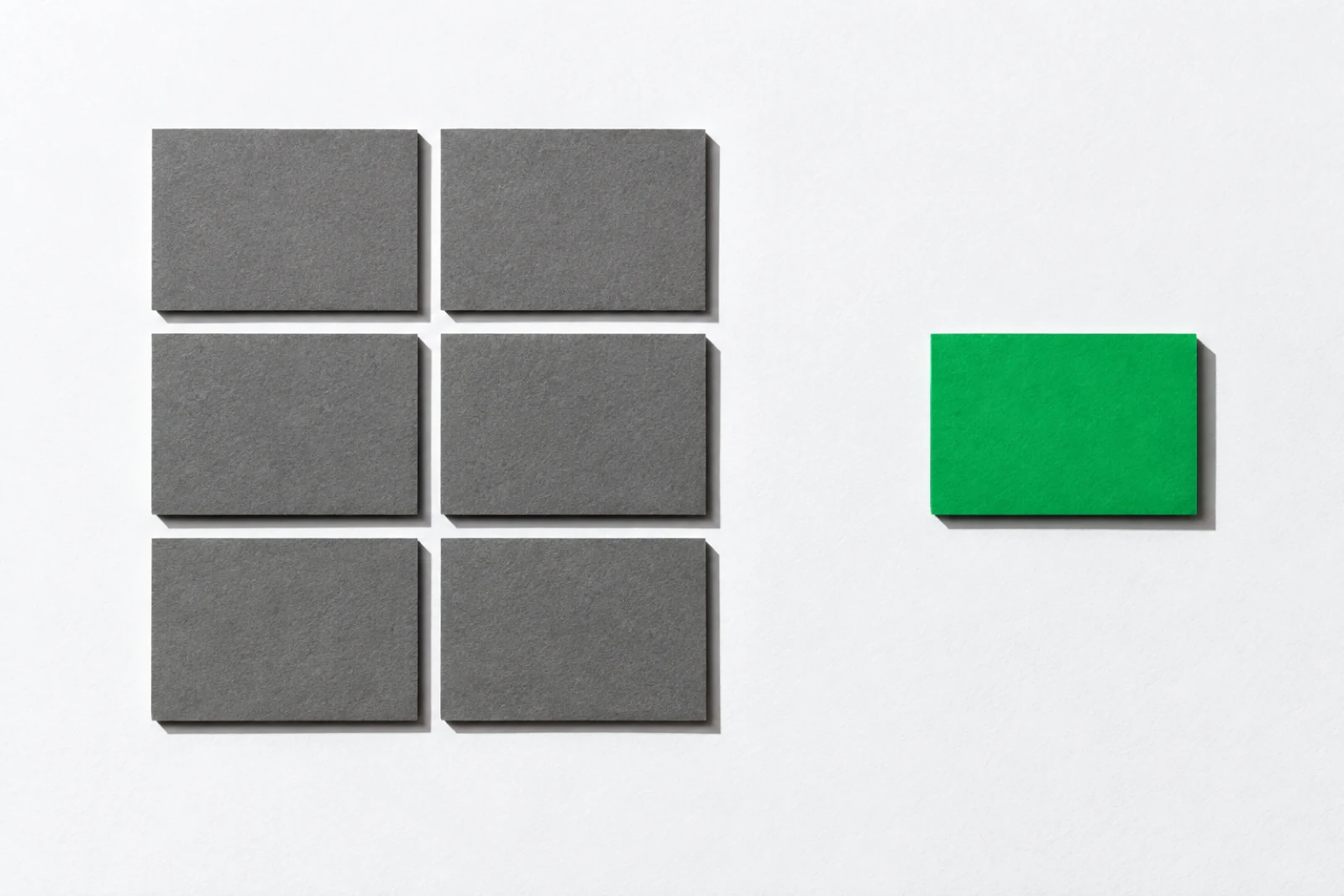 Six cartes de carton gris serrées en bloc de deux colonnes sur fond blanc, et une carte verte de même taille posée seule à l’écart sur la droite, ombres dures vers le bas à droite.