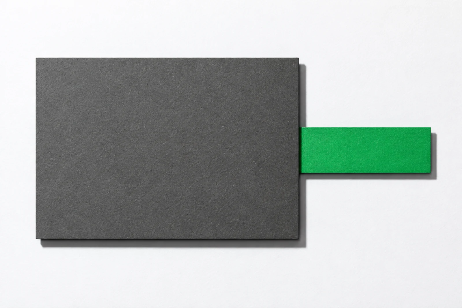 Grande plaque de carton gris posée à plat sur fond blanc, sous laquelle glisse une bande de papier verte qui dépasse franchement de son bord droit, ombres dures vers le bas à droite.
