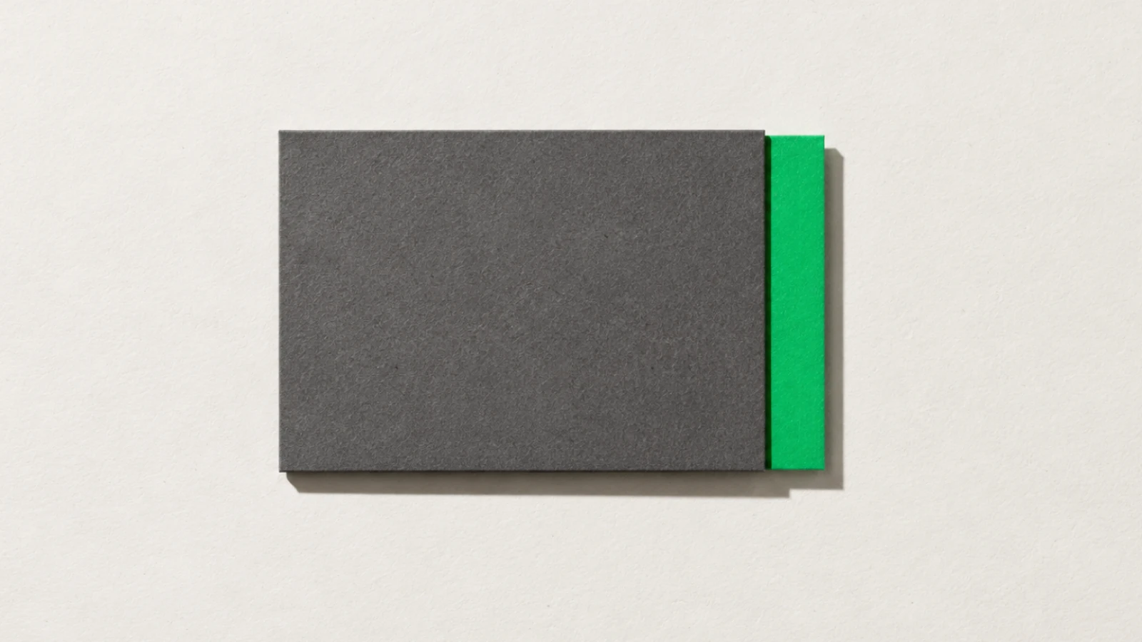 Carte de papier vert presque entièrement recouverte par une plaque de carton gris posée dessus, seule une bande verte dépasse sur la droite, ombre nette vers le bas à droite, sur fond beige.