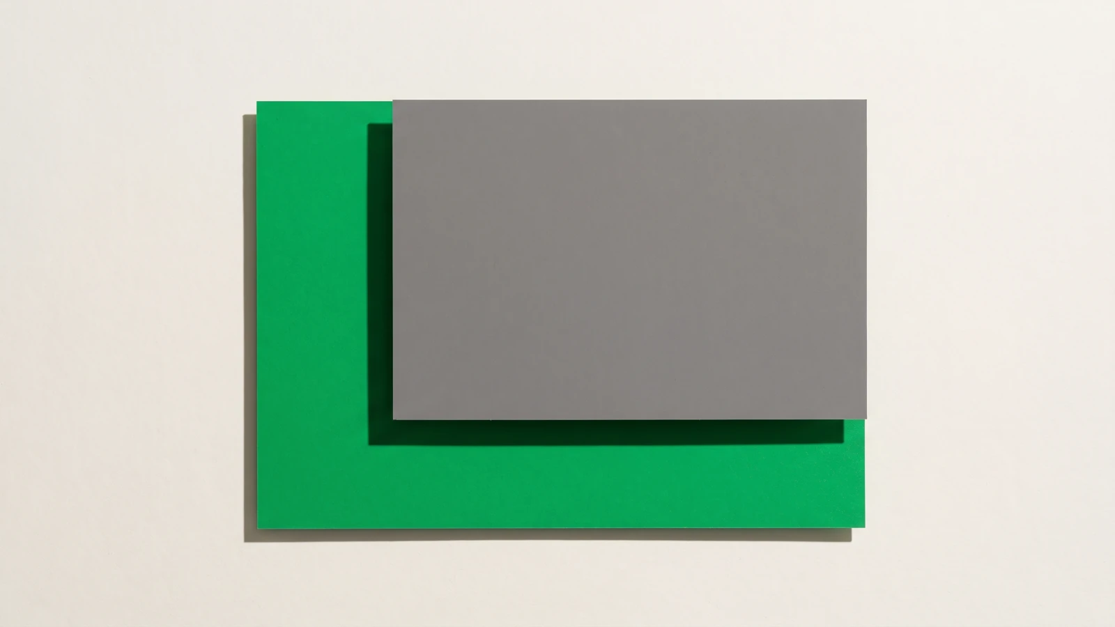 Une plaque de carton gris mat posée à plat sur une grande carte de papier vert dont elle recouvre plus de la moitié, ombres dures projetées vers le bas à droite