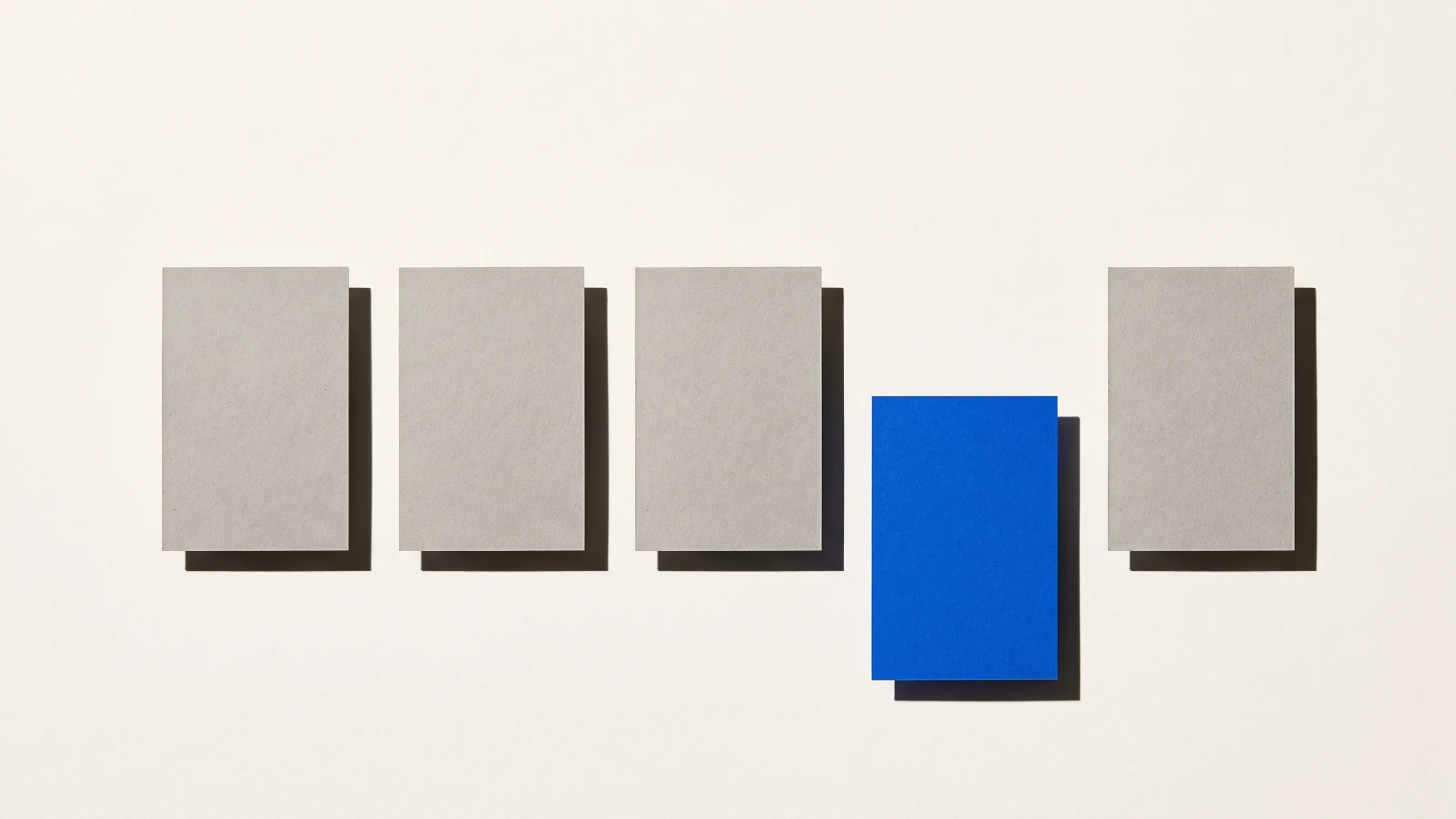 Cinq cartes alignées vues du dessus sur papier beige, quatre en carton gris et une bleue sortie de la rangée vers le bas, ombres dures projetées vers le bas à droite.