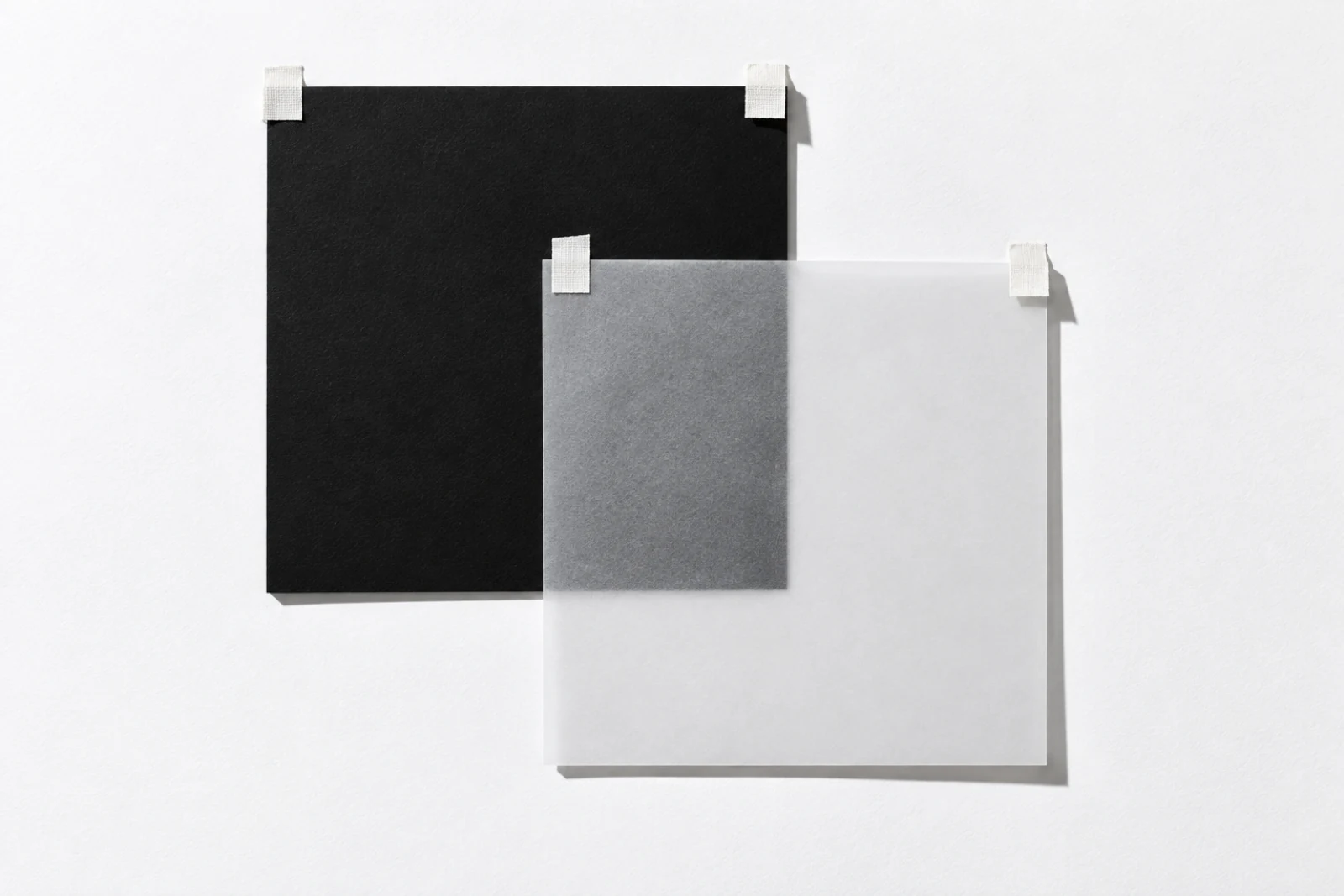 Vue de face, sur une surface blanche, un carré de papier noir mat et une feuille de papier calque de même taille scotchés en décalé vers le bas à droite ; le coin du noir vu à travers le calque devient gris ; ombres dures vers le bas à droite.