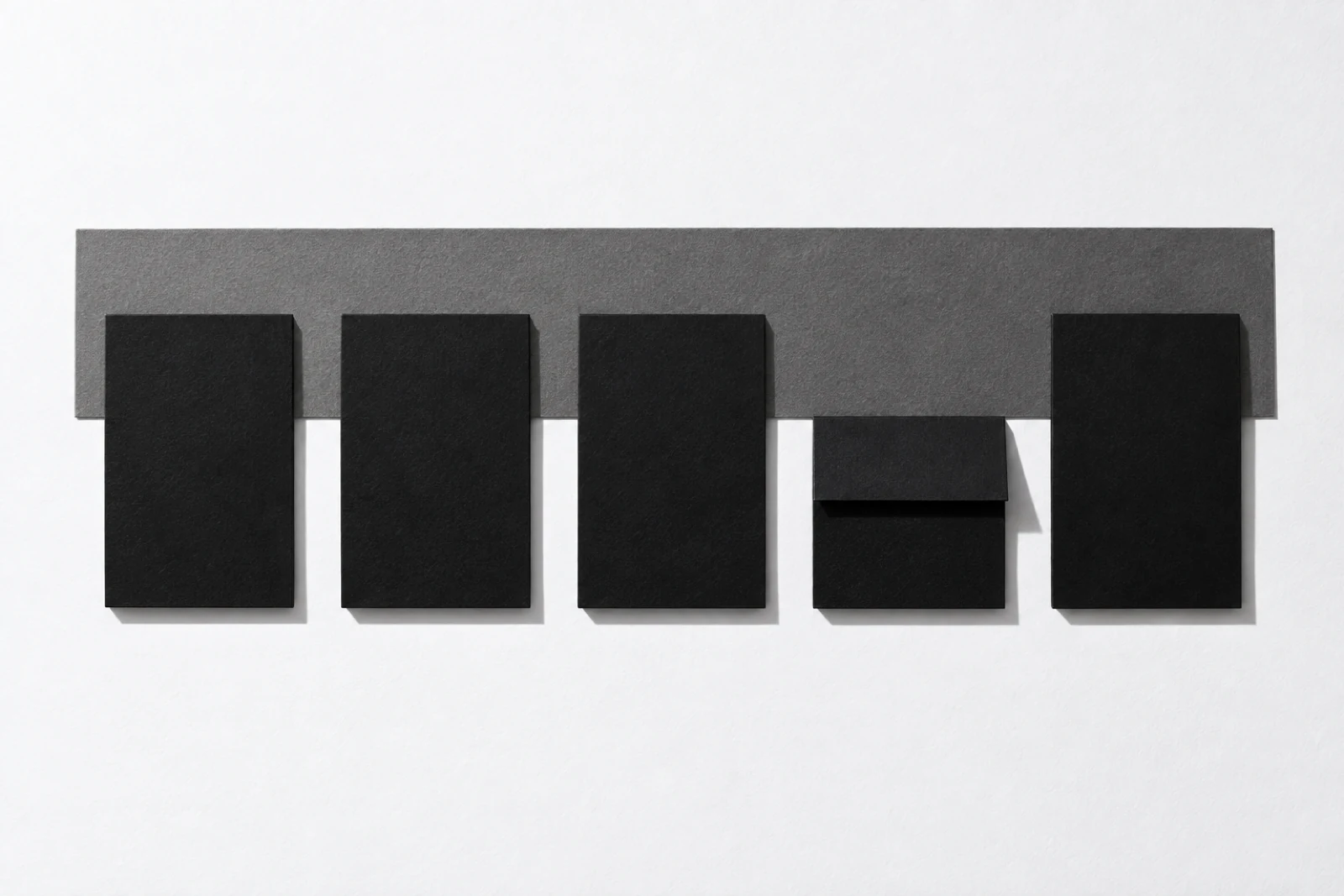 Vue de dessus, sur un fond blanc, cinq cartes de papier noir mat alignées sur une bande de carton gris ; la quatrième, pliée en deux, est deux fois plus courte que les autres ; ombres dures vers le bas à droite.