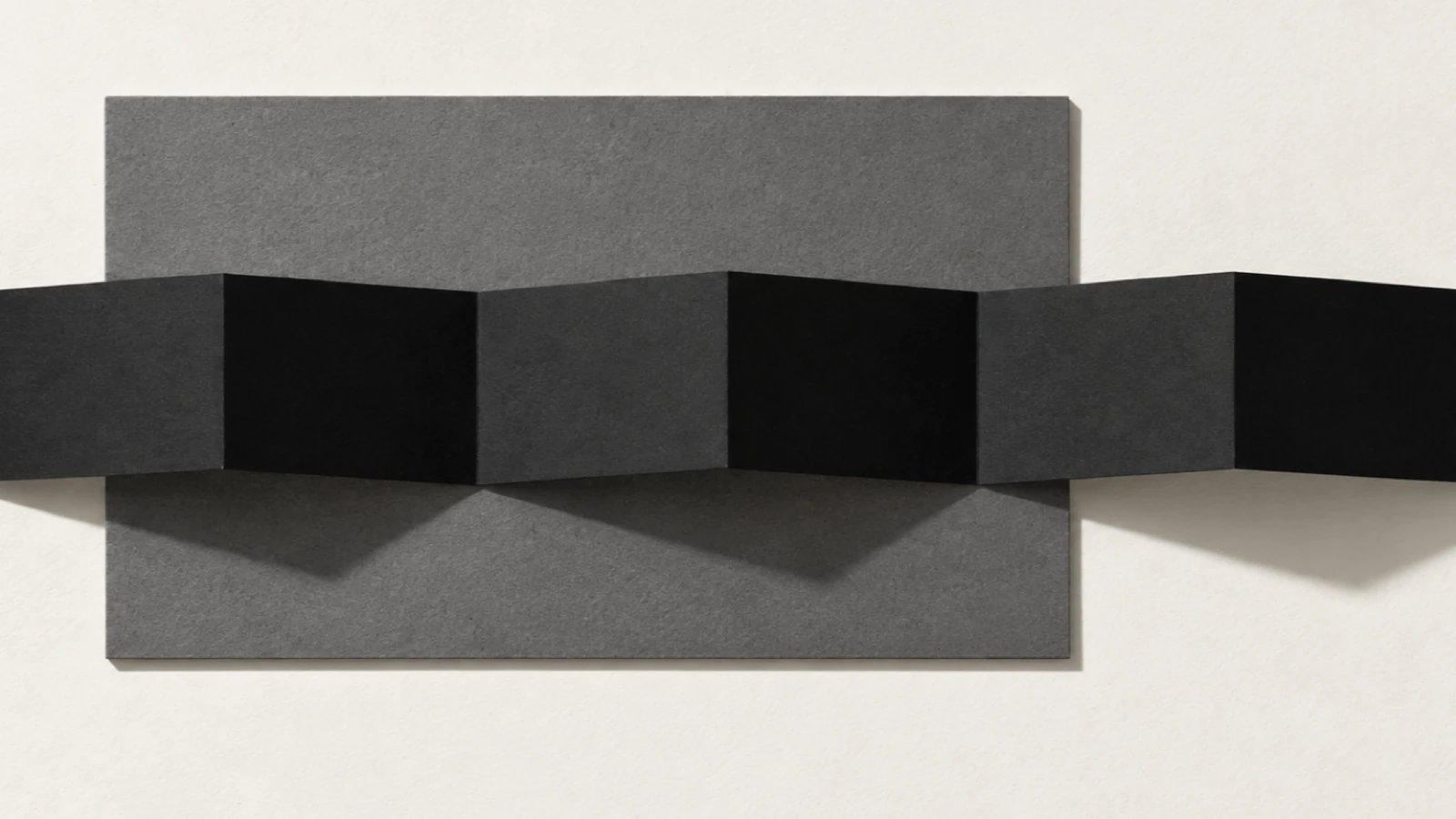 Vue de dessus, sur un fond de papier beige, une longue bande de papier noir mat pliée en accordéon traverse une plaque de carton gris ; les pans éclairés et les pans dans l’ombre alternent, ombres dures vers le bas.