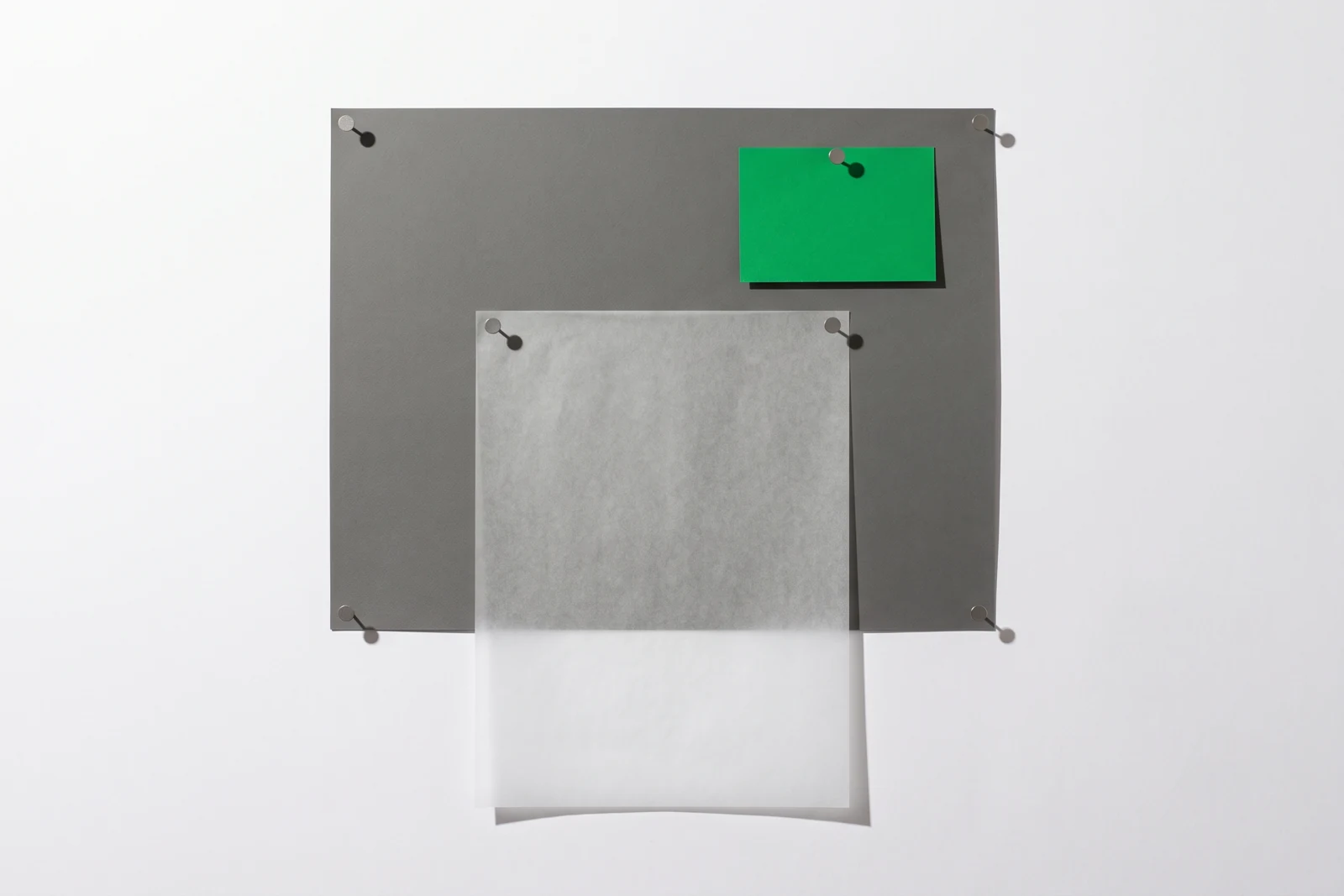Un grand carton gris punaisé à plat sur fond blanc, recouvert aux deux tiers par une feuille de calque punaisée en haut, et un petit rectangle de papier vert punaisé dans la zone restée nue