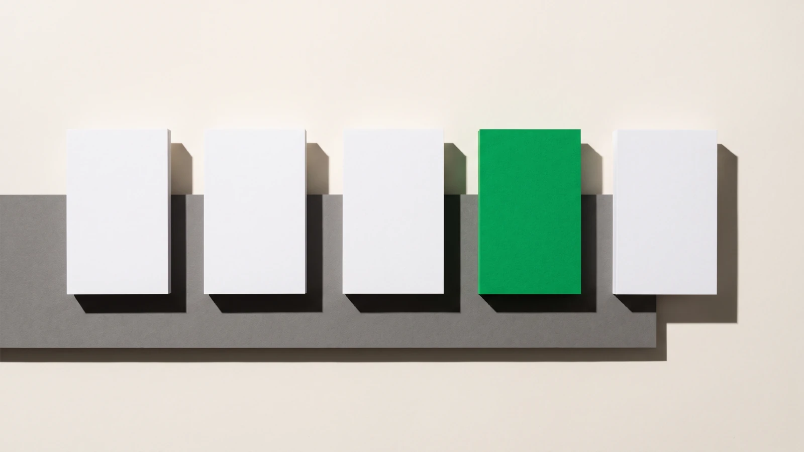 Cinq cartes de papier alignées à pas régulier sur une bande de carton gris, quatre blanches et une verte à la quatrième place, sur fond papier beige, une seule ombre nette vers le bas à droite