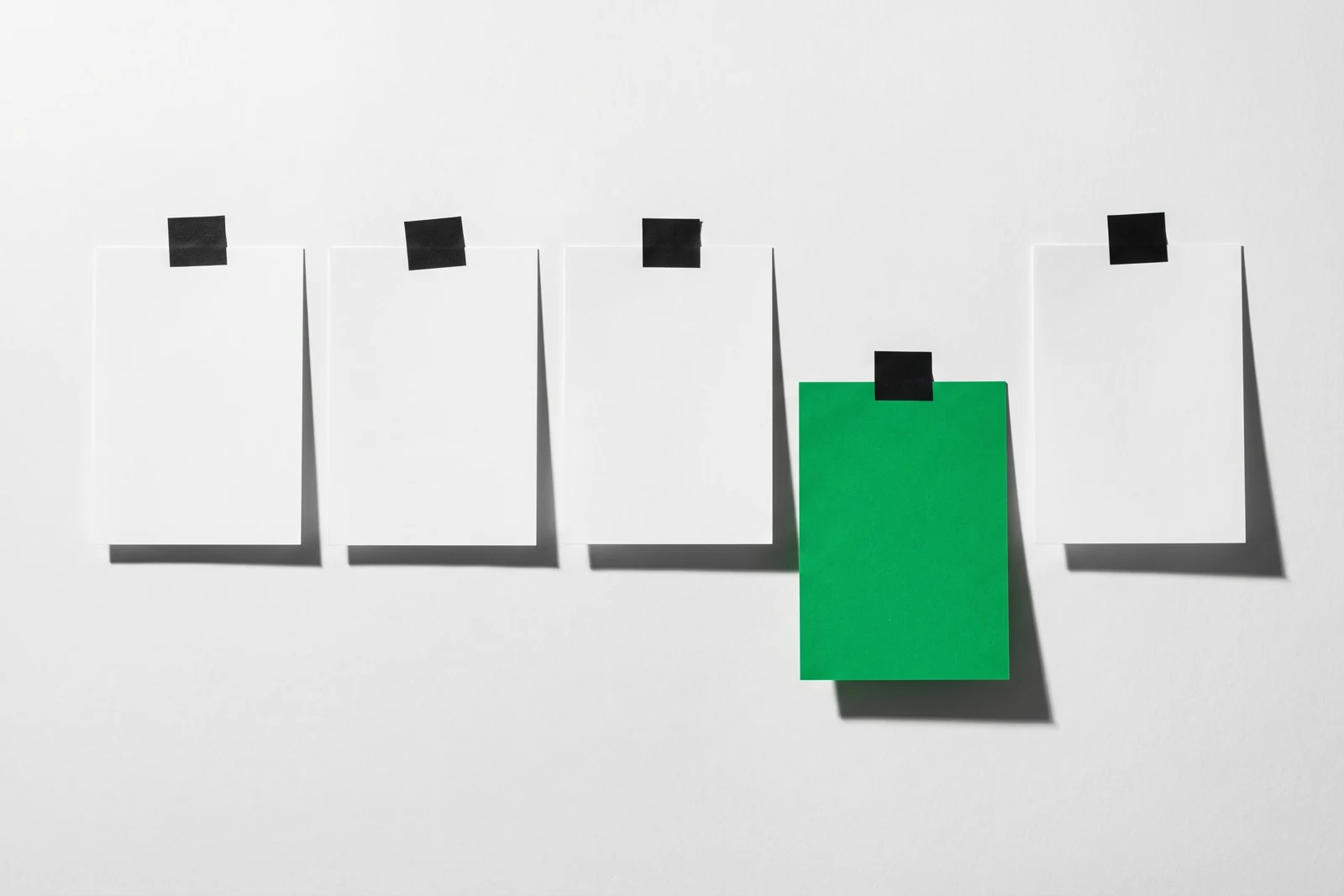 Cinq feuilles de papier scotchées en ligne sur un mur blanc, la quatrième, verte, décalée vers le bas d'une demi-hauteur, ombres dures vers le bas à droite.