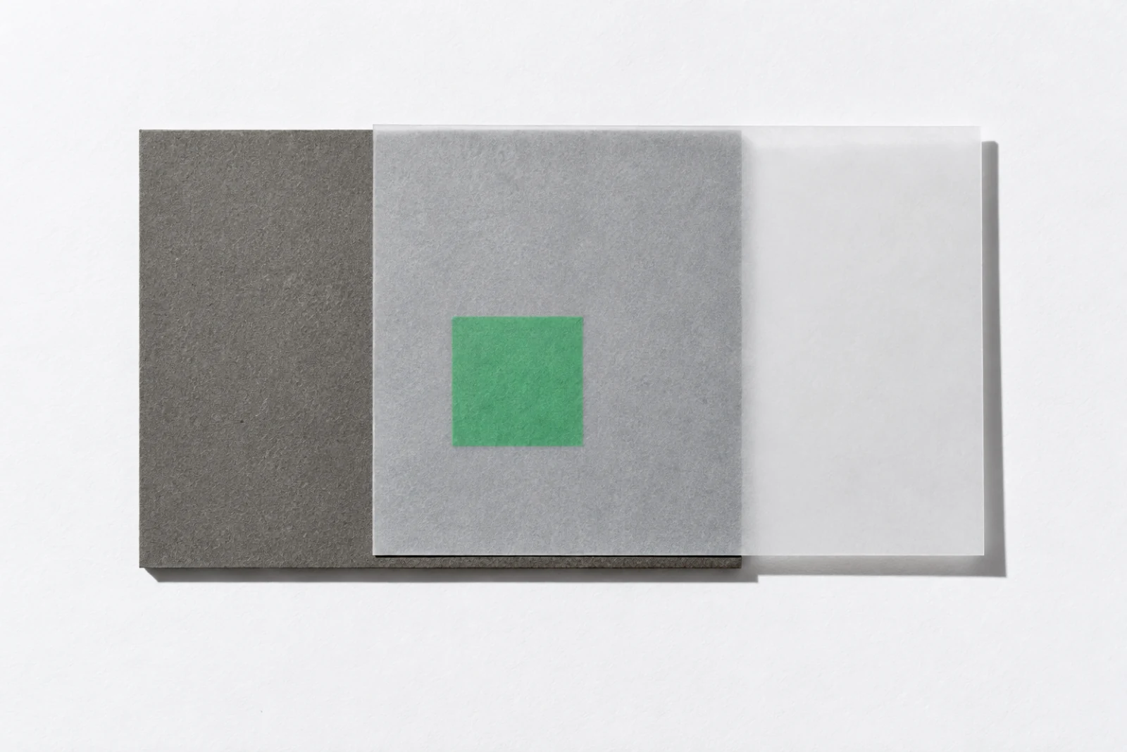 Une feuille de papier calque posée en décalage sur un carton gris, laissant voir par transparence un petit carré vert glissé dessous.