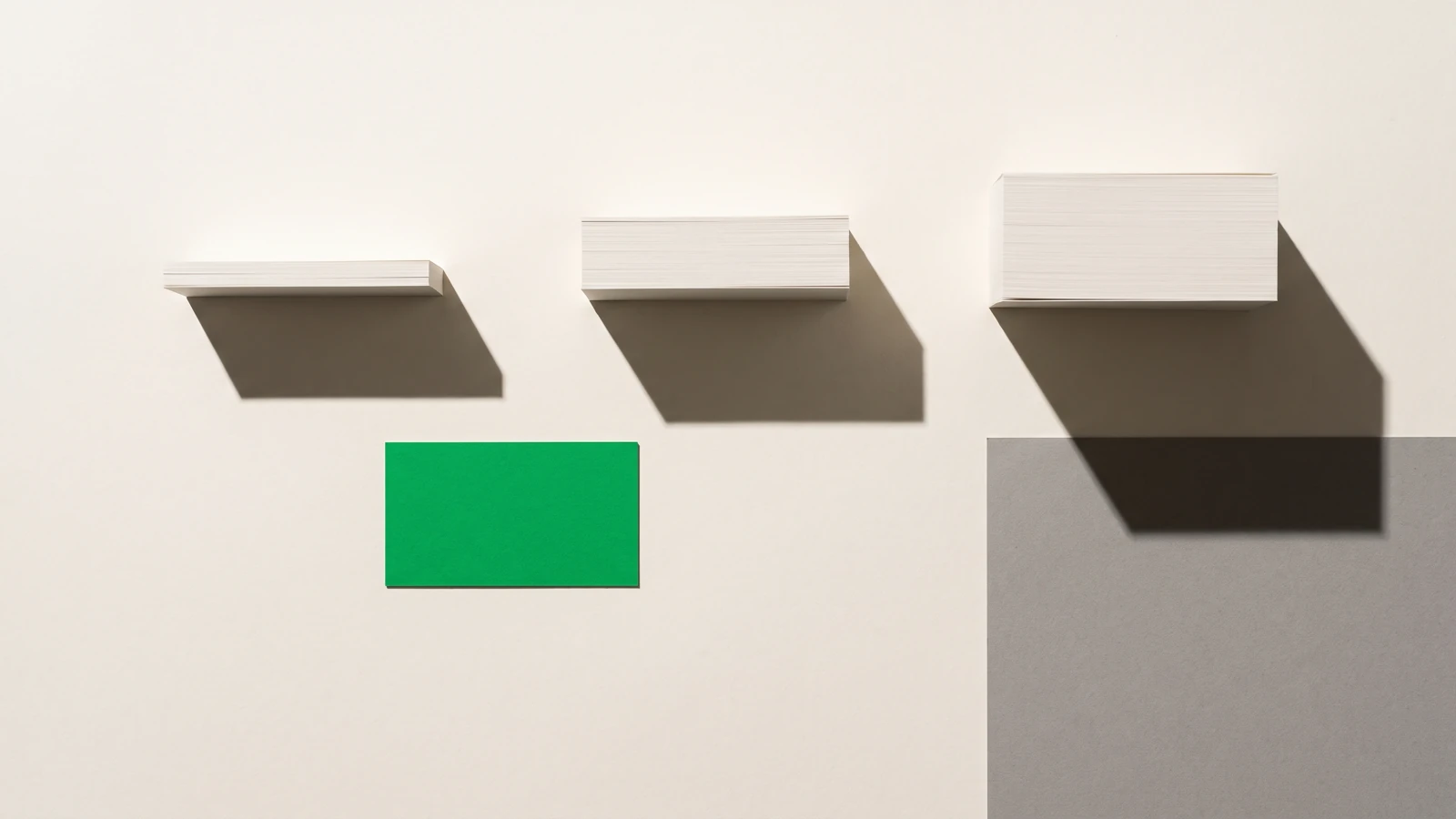 Des piles de papier vierge d’épaisseurs nettement inégales alignées sur une même base, ombres dures projetées vers le bas à droite.