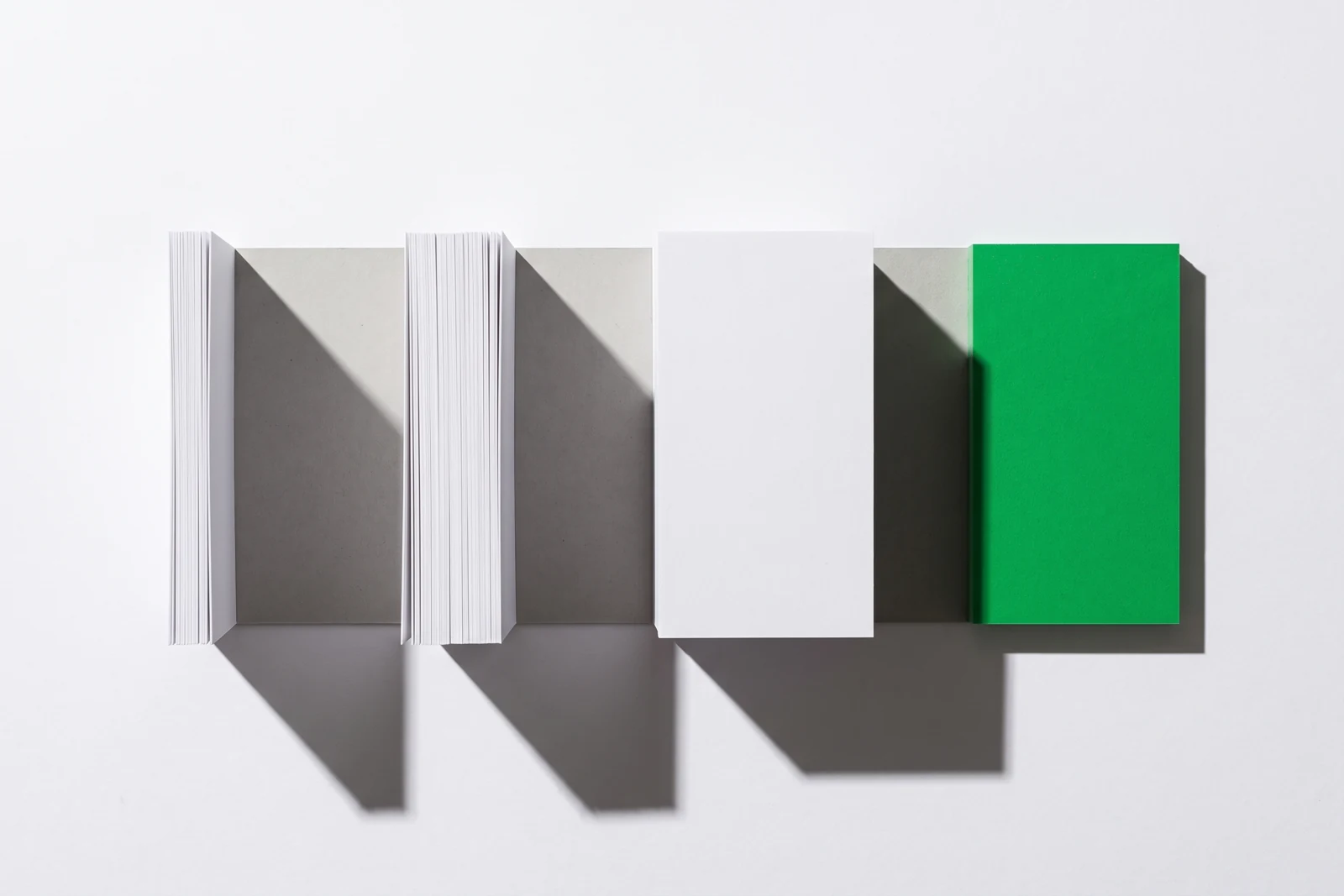 Des piles de papier vierge d’épaisseurs nettement inégales alignées sur une même base, ombres dures projetées vers le bas à droite.