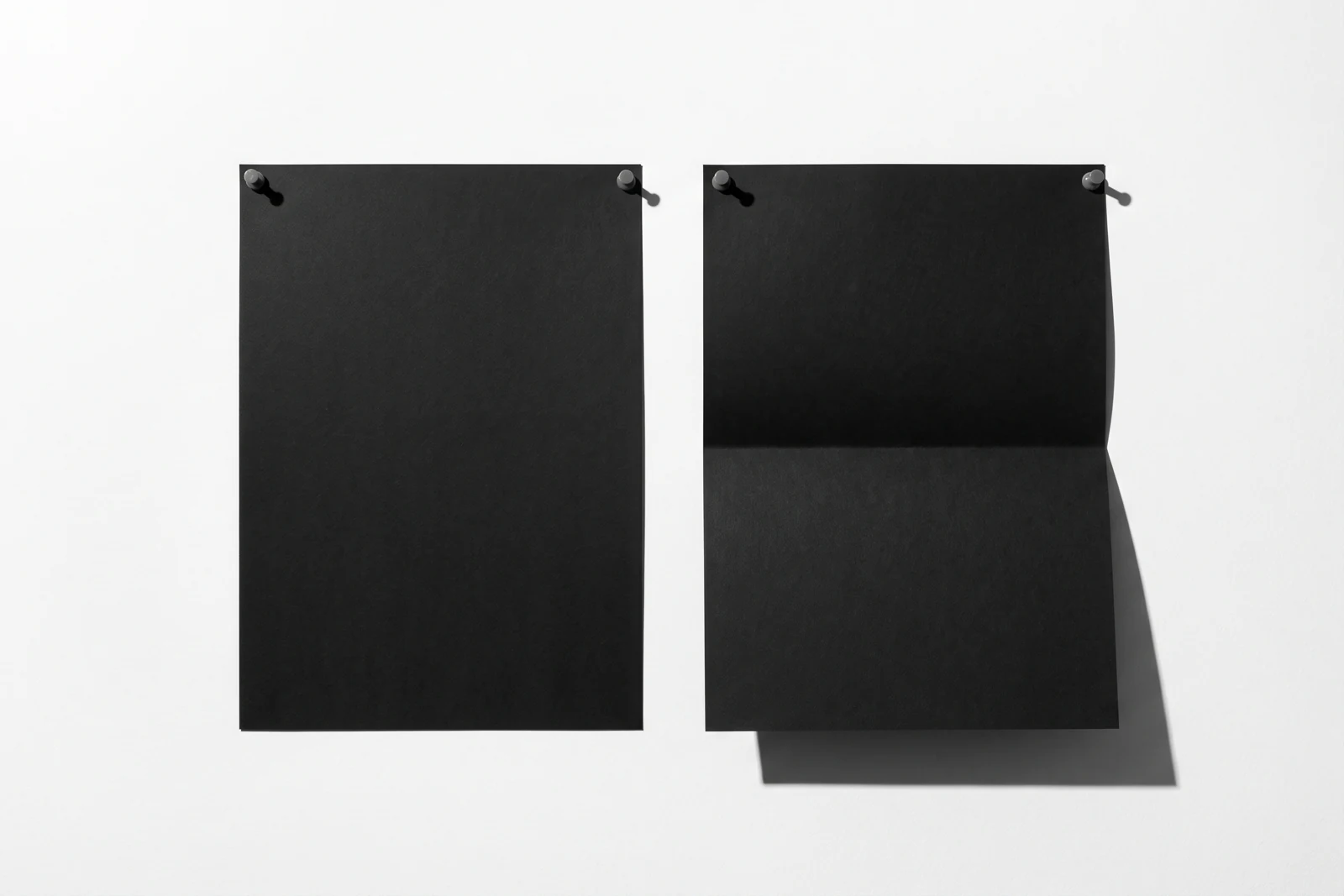 Vue de face, sur une surface verticale blanche qui remplit tout le cadre, deux feuilles de papier noir mat de même format, punaisées côte à côte ; celle de droite est pliée en deux, sa moitié basse rabattue vers l’avant ; ombres dures vers le bas à droite.