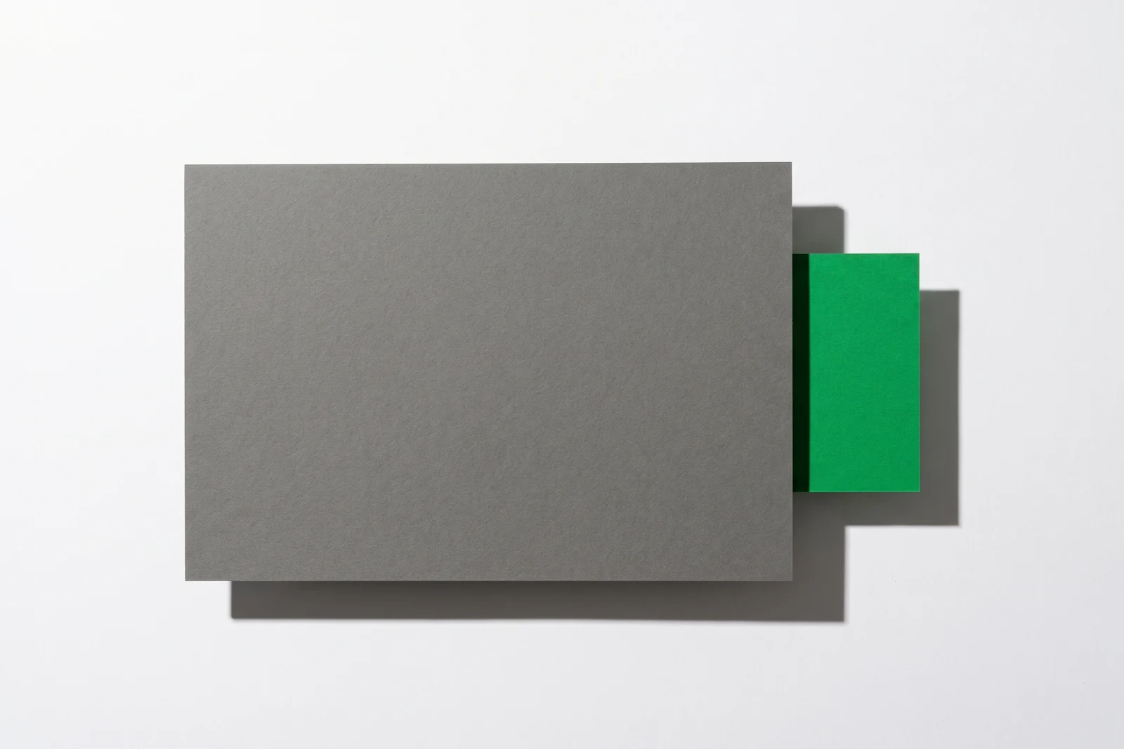 Une grande feuille de carton gris posée à plat sur fond blanc, une carte verte glissée dessous qui dépasse seulement par le bord droit, ombres dures vers le bas à droite.