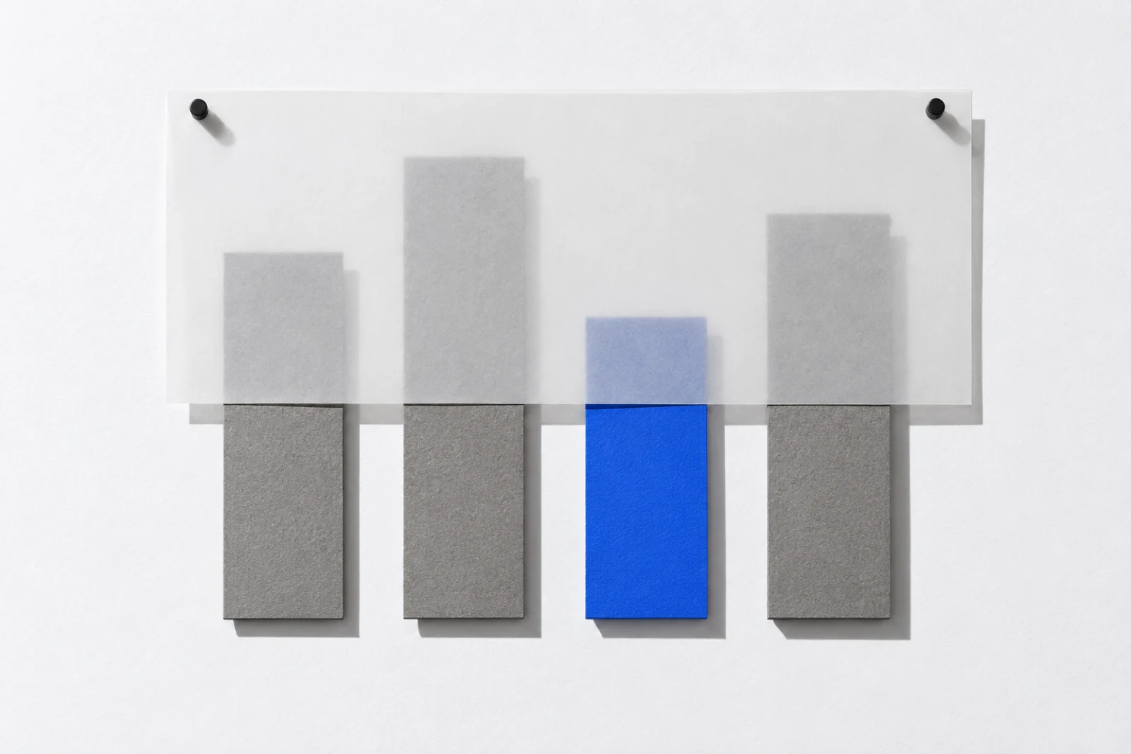 Vue de face, sur une surface blanche, quatre bandes de hauteurs inégales punaisées côte à côte, trois en carton gris et une en papier bleu ; une feuille de papier calque couvre le haut de la rangée ; ombres dures vers le bas à droite.