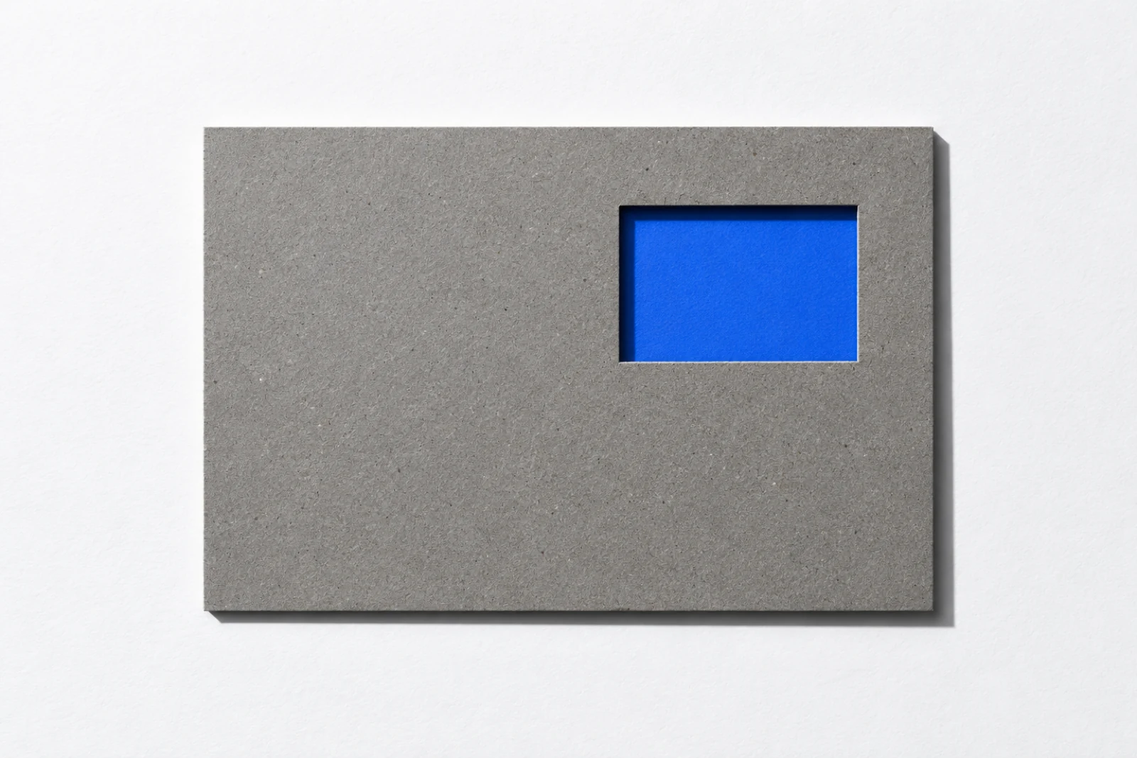 Vue de dessus, sur un fond blanc, une plaque de carton gris percée d’une fenêtre rectangulaire en haut à droite, par laquelle on voit une feuille de papier bleu posée dessous ; ombres dures vers le bas à droite.