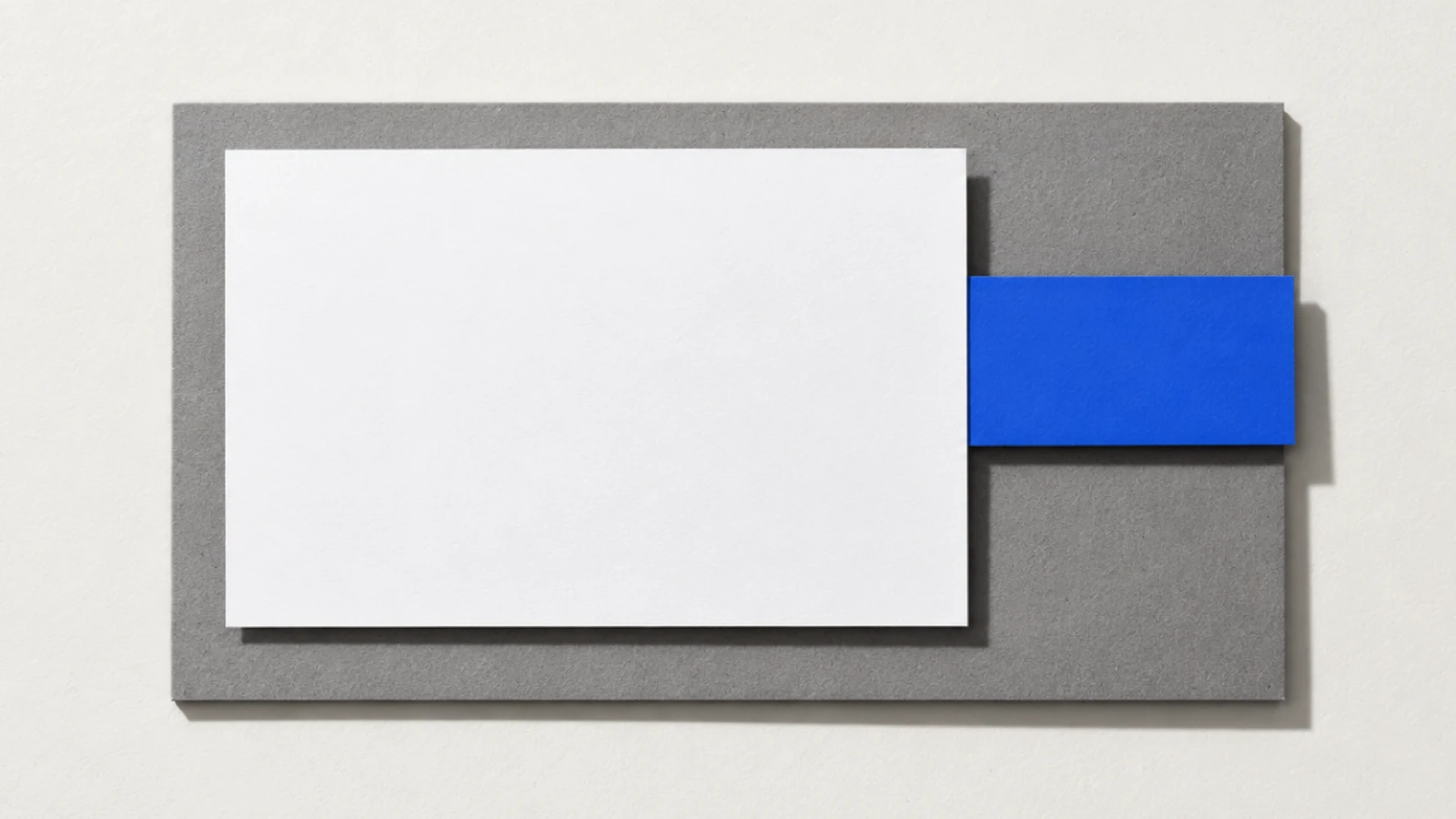 Vue de dessus, sur un fond de papier beige, une feuille blanche posée sur une plaque de carton gris ; une bande de papier bleu glissée sous la feuille dépasse nettement sur la droite ; ombres dures vers le bas à droite.
