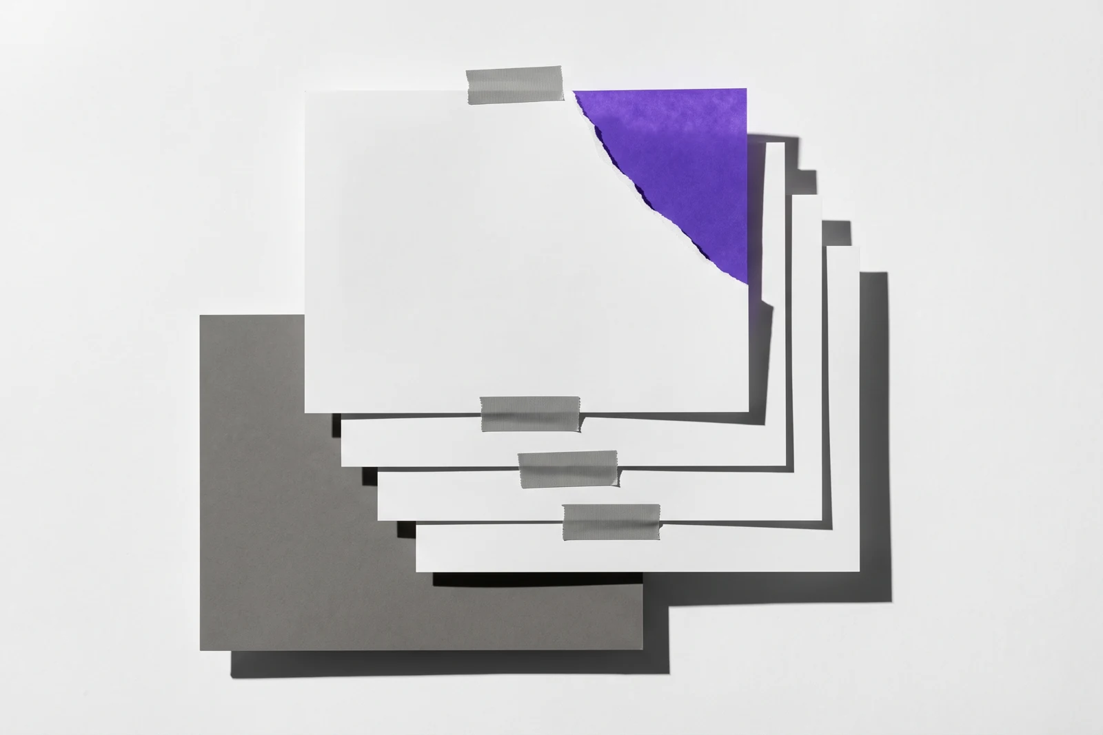Des feuilles de papier scotchées à plat, le coin de la feuille du dessus arraché, ombres dures projetées vers le bas à droite.