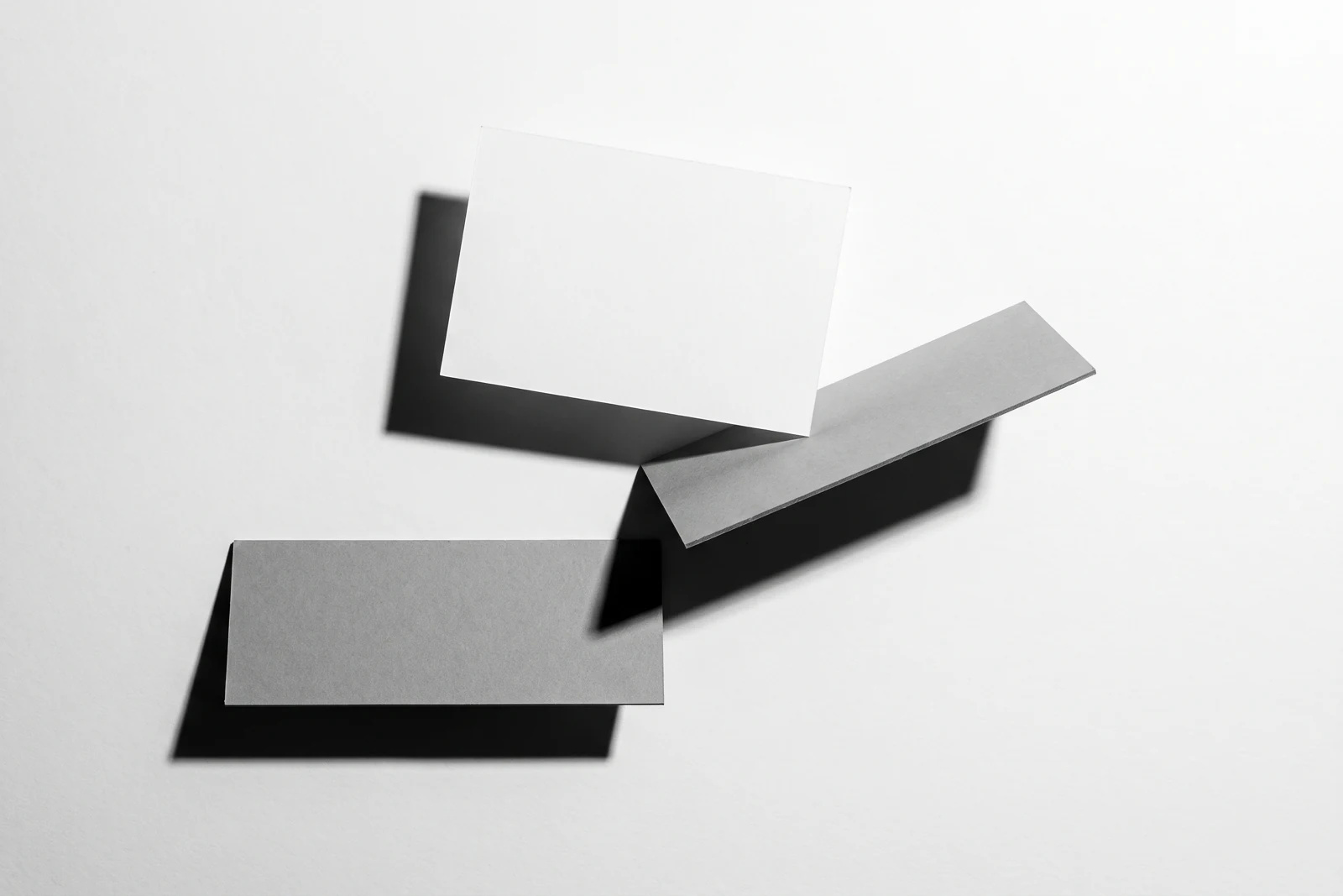 Une carte de papier en équilibre sur le bord d’une plaque de carton gris, ombres dures projetées vers le bas à droite.