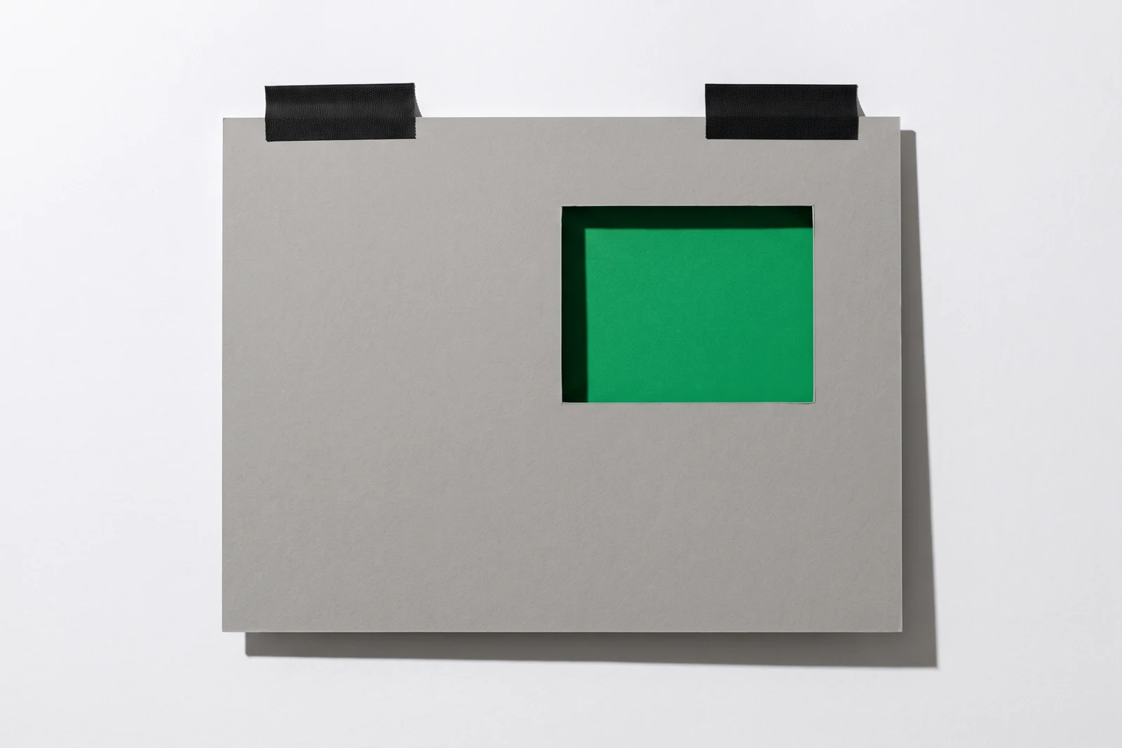 Une plaque de carton gris scotchée au mur par deux bandes de toile noire, percée d'une fenêtre rectangulaire qui laisse voir un papier vert placé derrière.