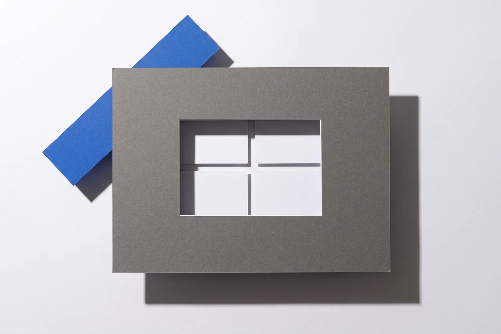Une fenêtre rectangulaire découpée dans une plaque de carton gris laisse voir des cartes dessous, ombres dures projetées vers le bas à droite.