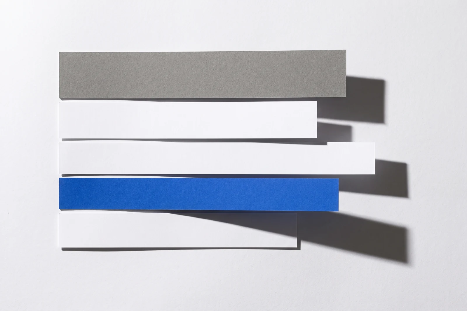 Des bandes de papier vierge alignées à fleur sur leur bord gauche, de longueurs différentes, ombres dures projetées vers le bas à droite.
