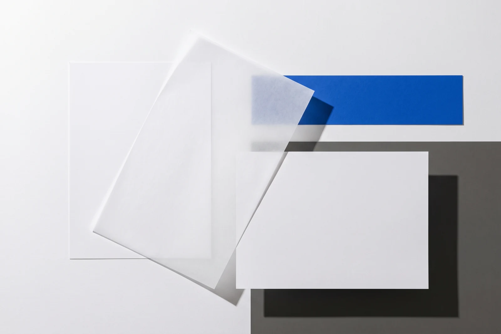 Un calque cristal posé de biais sur des feuilles de papier vierge, ombres dures projetées vers le bas à droite.